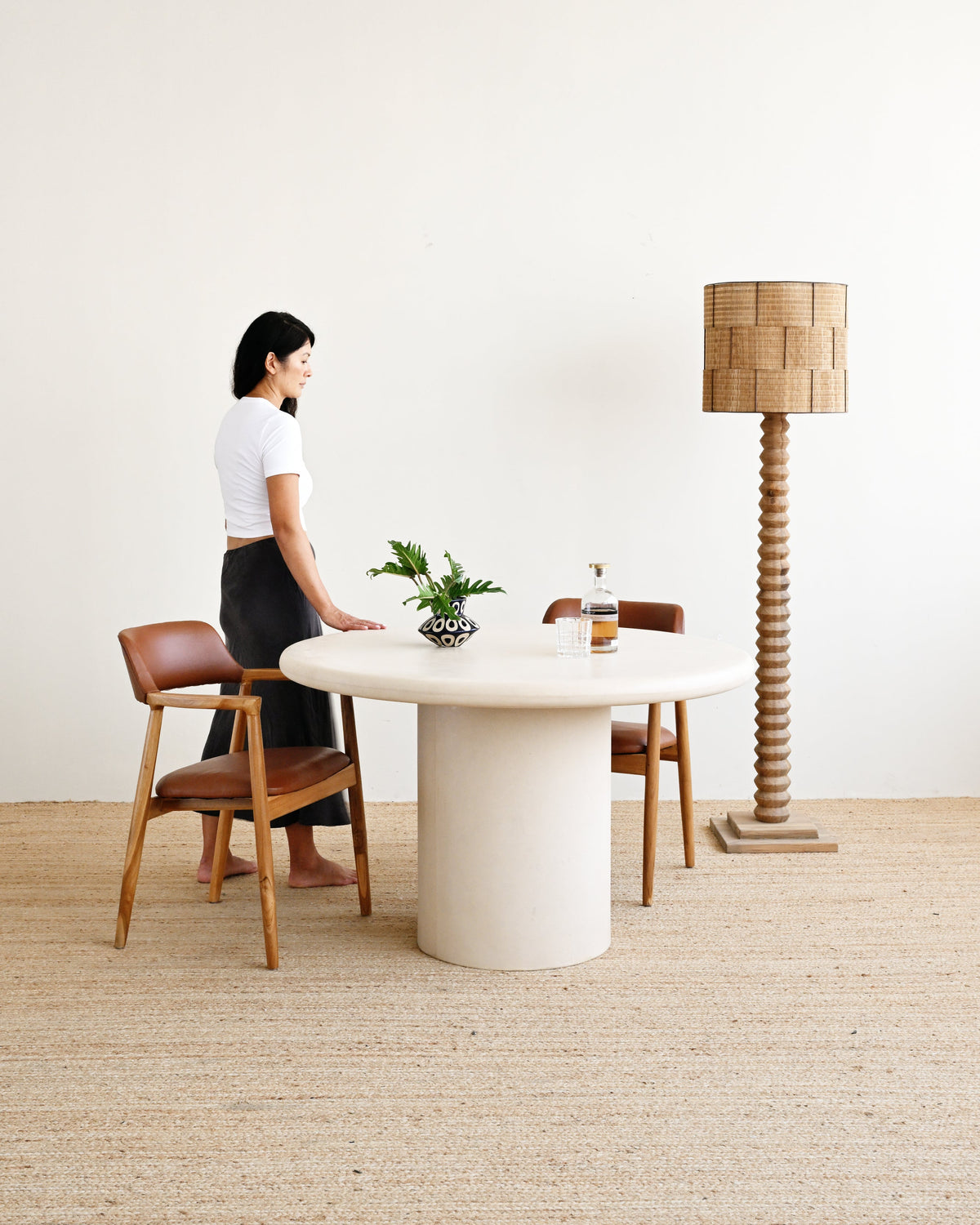 A woman stands beside a round off-white pedestal dining table with two mid-century style wooden chairs with brown leather seats, in a minimalist room with a woven floor and a tall woven lamp.