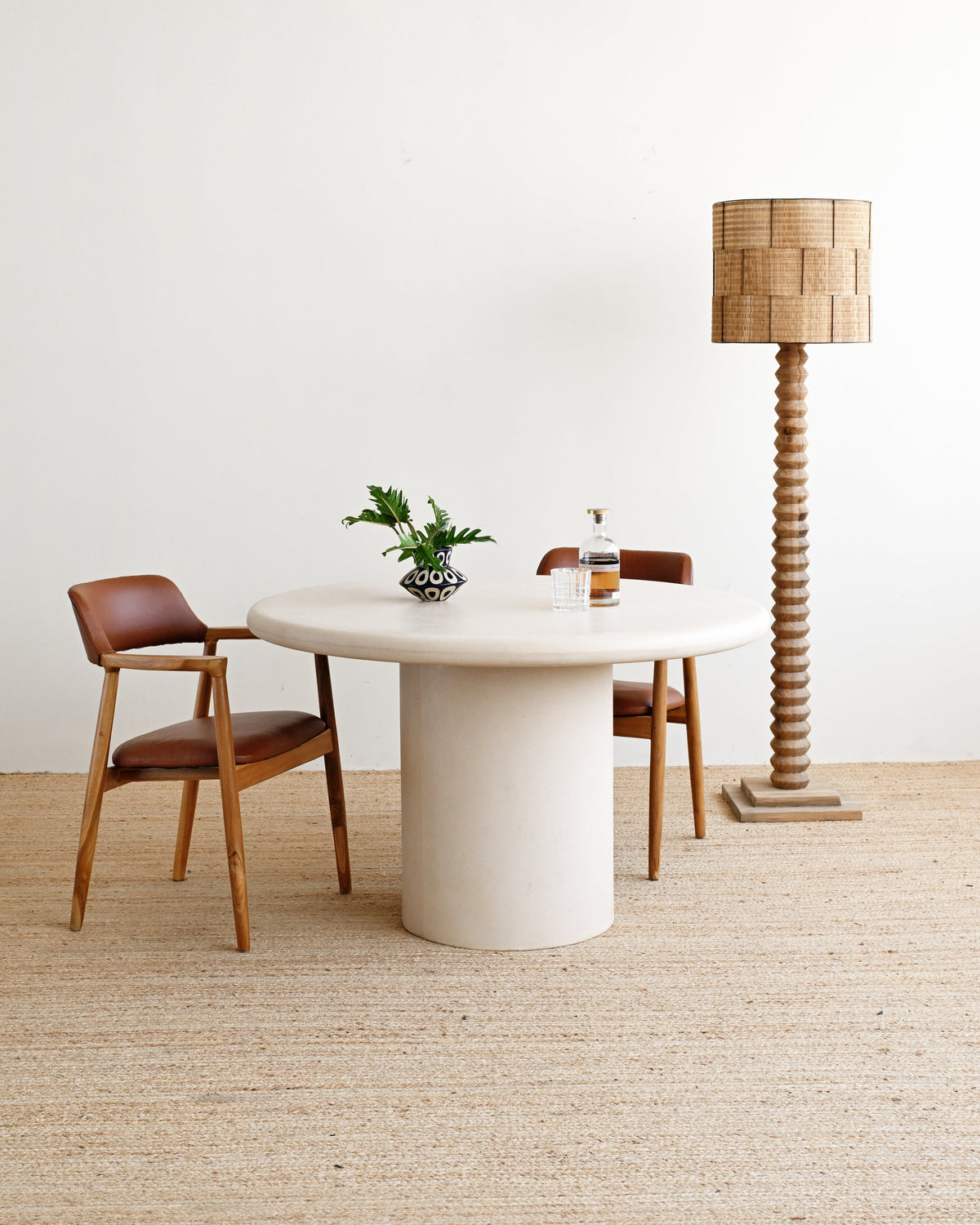 A minimalist dining setup featuring a round white stone table with a thick pedestal base, accompanied by two mid-century modern wooden chairs with brown leather seats, a tall woven wood floor lamp, and a small potted green plant on a light beige jute rug against a white wall.