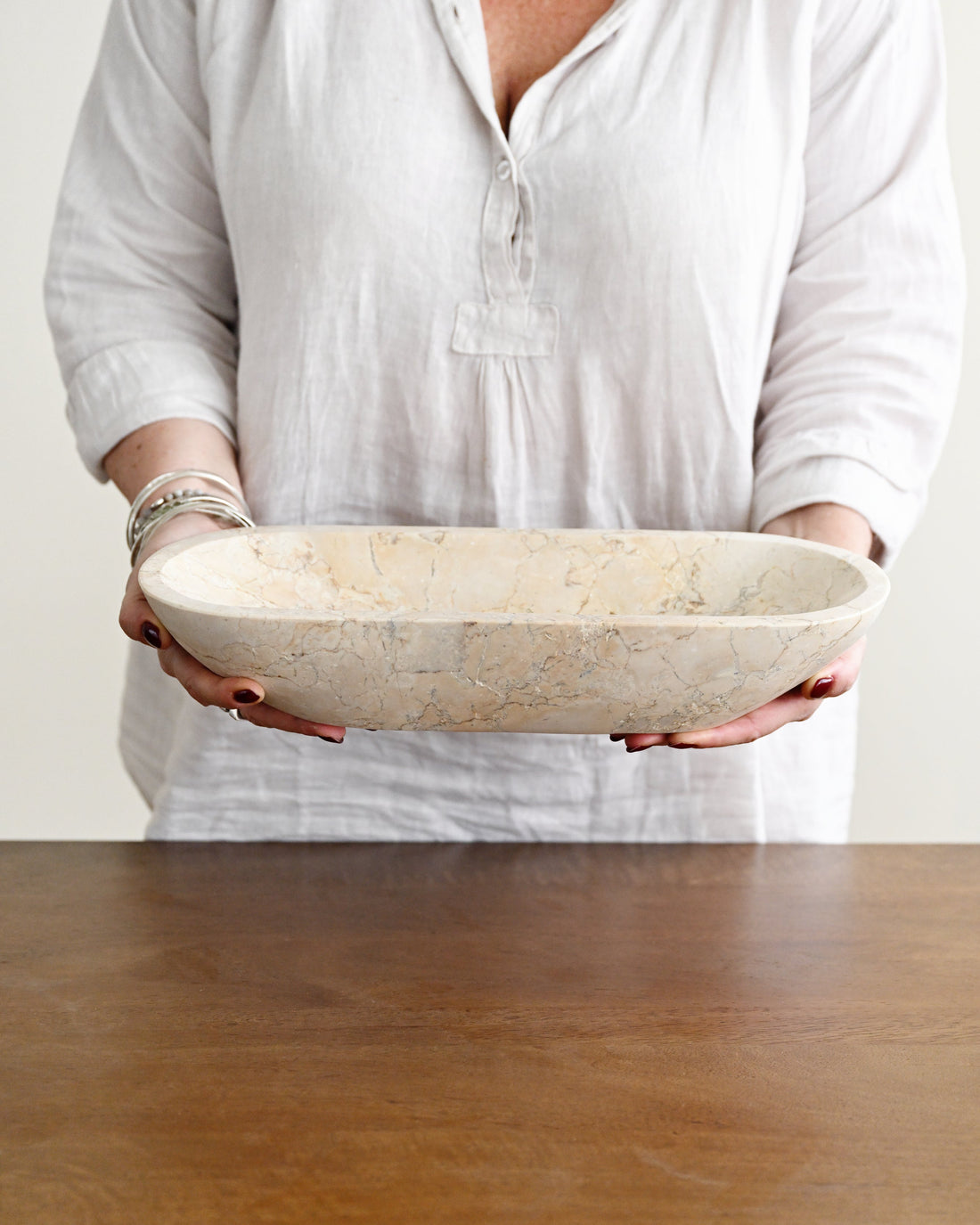 A person in a white linen shirt holds a large, oval stone bowl with a textured beige and cream surface over a wooden table.