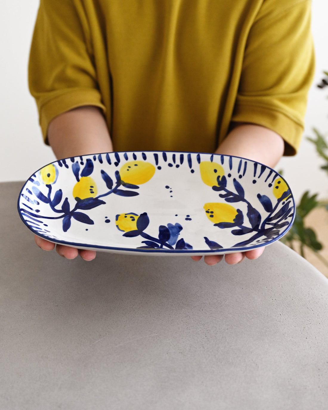 A person in a mustard yellow shirt holds a white oval serving platter decorated with a blue and yellow lemon pattern.