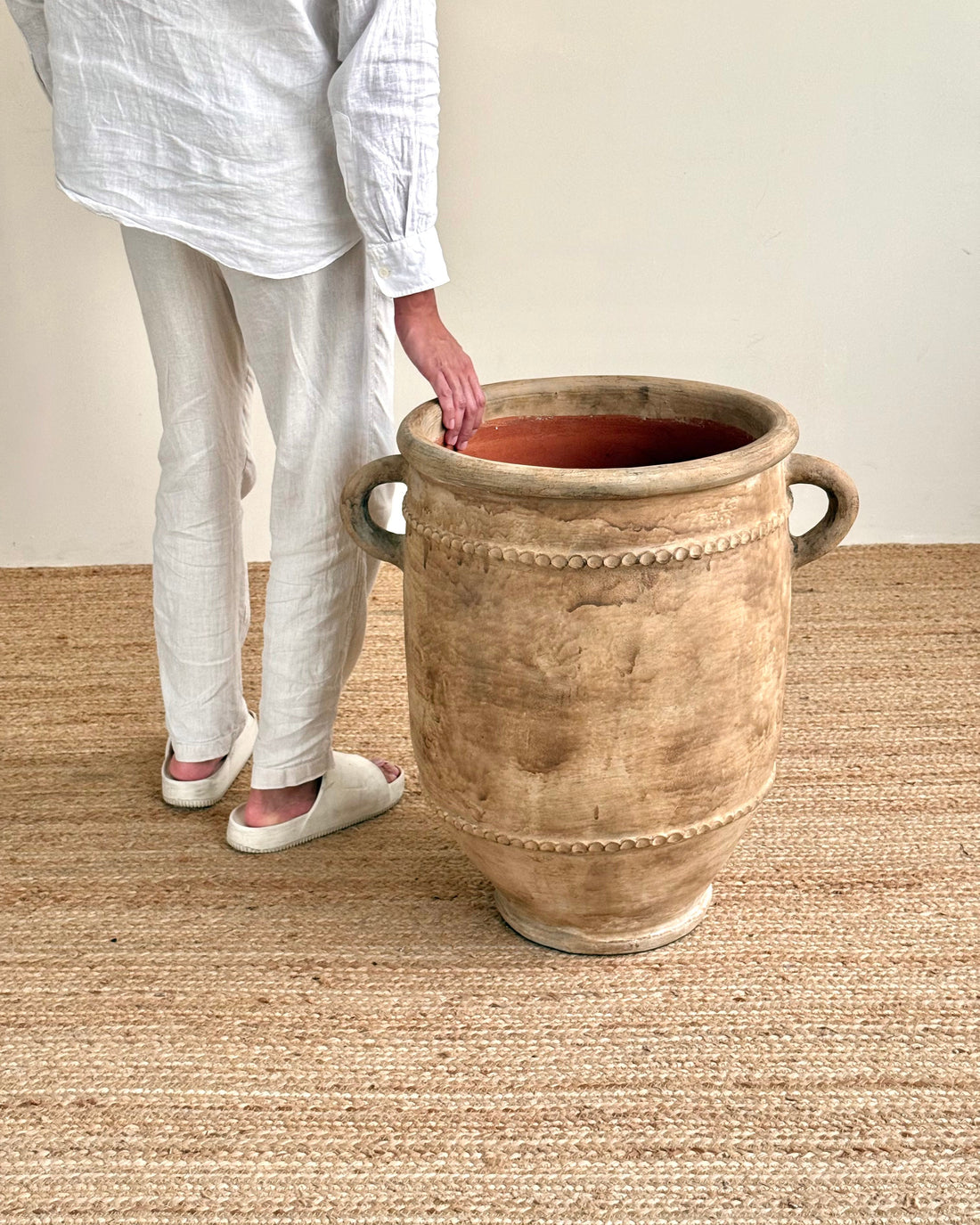 A person wearing a white linen shirt and pants stands beside a large rustic beige ceramic planter pot with side handles on a woven jute rug.