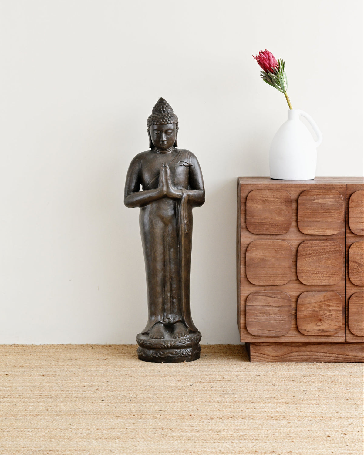 A dark bronze standing Buddha statue displayed on a light beige woven rug next to a wooden cabinet with a white vase holding a pink flower, set against a plain off-white wall.