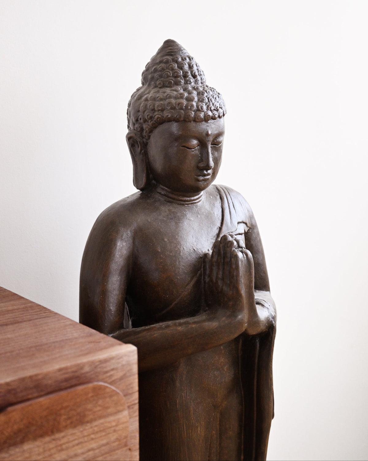A dark bronze Buddha statue with hands in a prayer pose, shown standing next to a wooden surface against a plain off-white background.