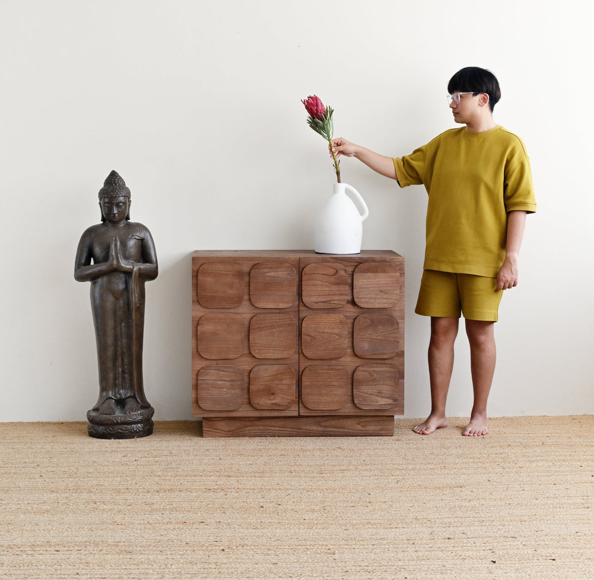 A child wearing a mustard yellow t-shirt and matching shorts stands barefoot next to a carved wooden chest, holding a white vase with a red flower, in a minimalist room with a Buddha statue and a woven rug.