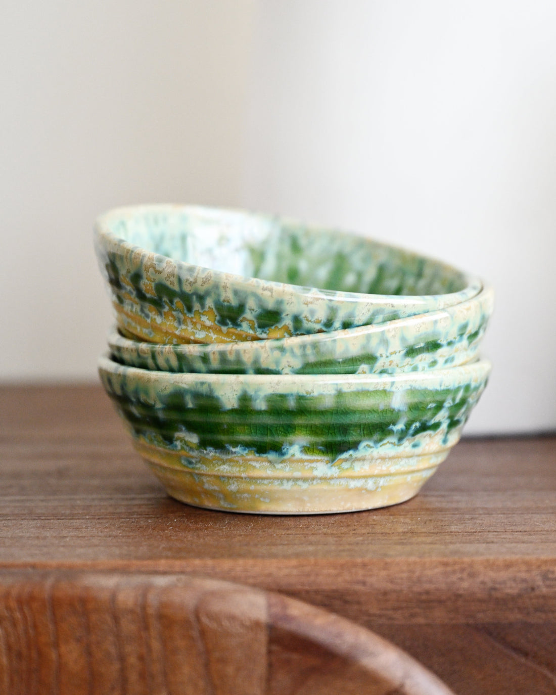 Three small stacked ceramic bowls with a green and yellow marbled glaze, resting on a wooden surface against a soft off-white background.