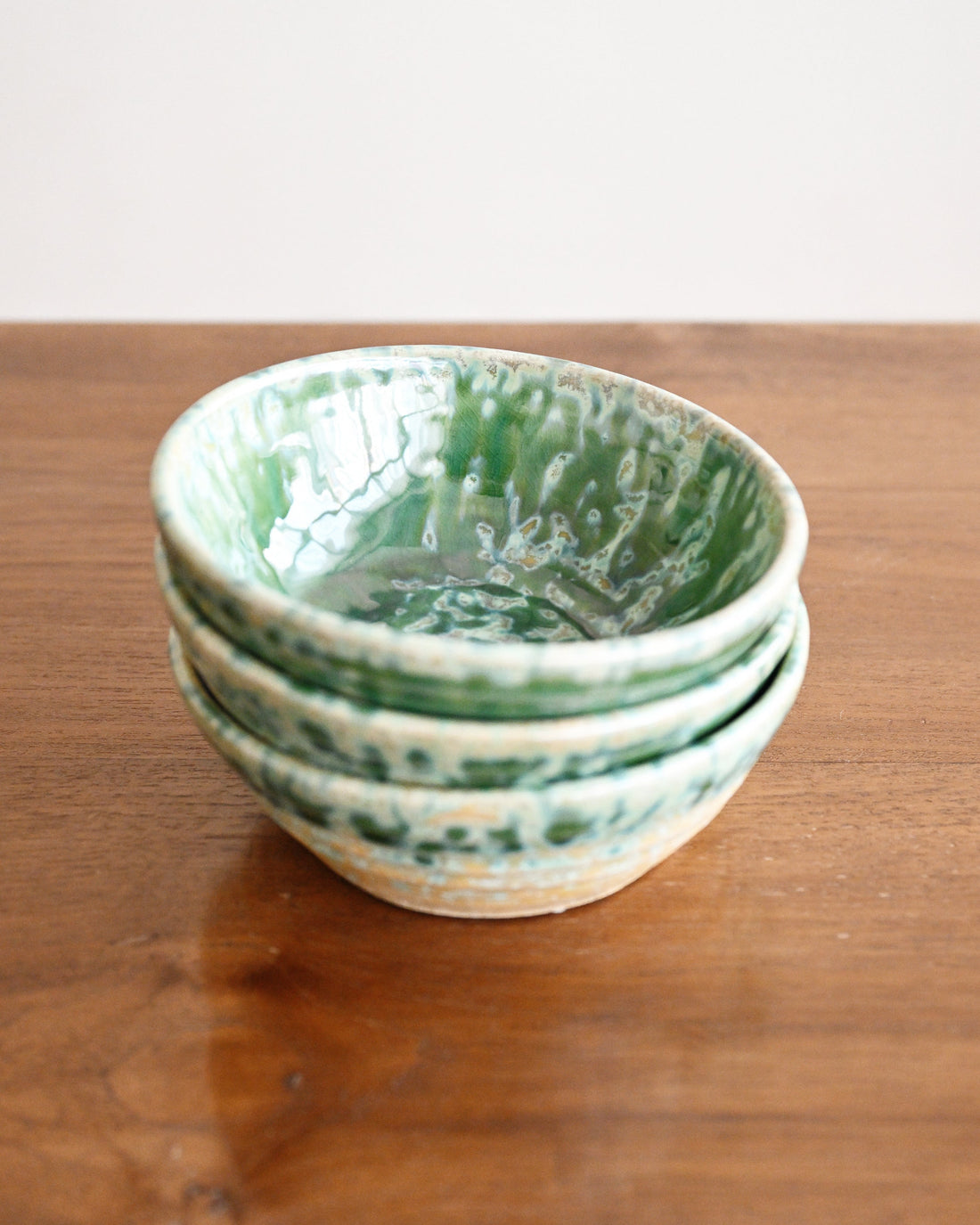 A stack of four handmade ceramic bowls with a green and white marbled glaze, displayed on a wooden tabletop against a neutral off-white background.