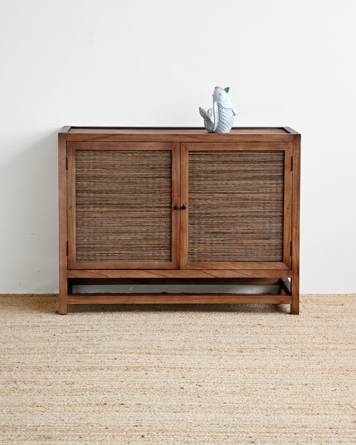 A mid-century modern wooden credenza or sideboard cabinet with two woven rattan doors, shown on a textured jute rug against a plain white wall. A small gray ceramic bird figurine sits on top.