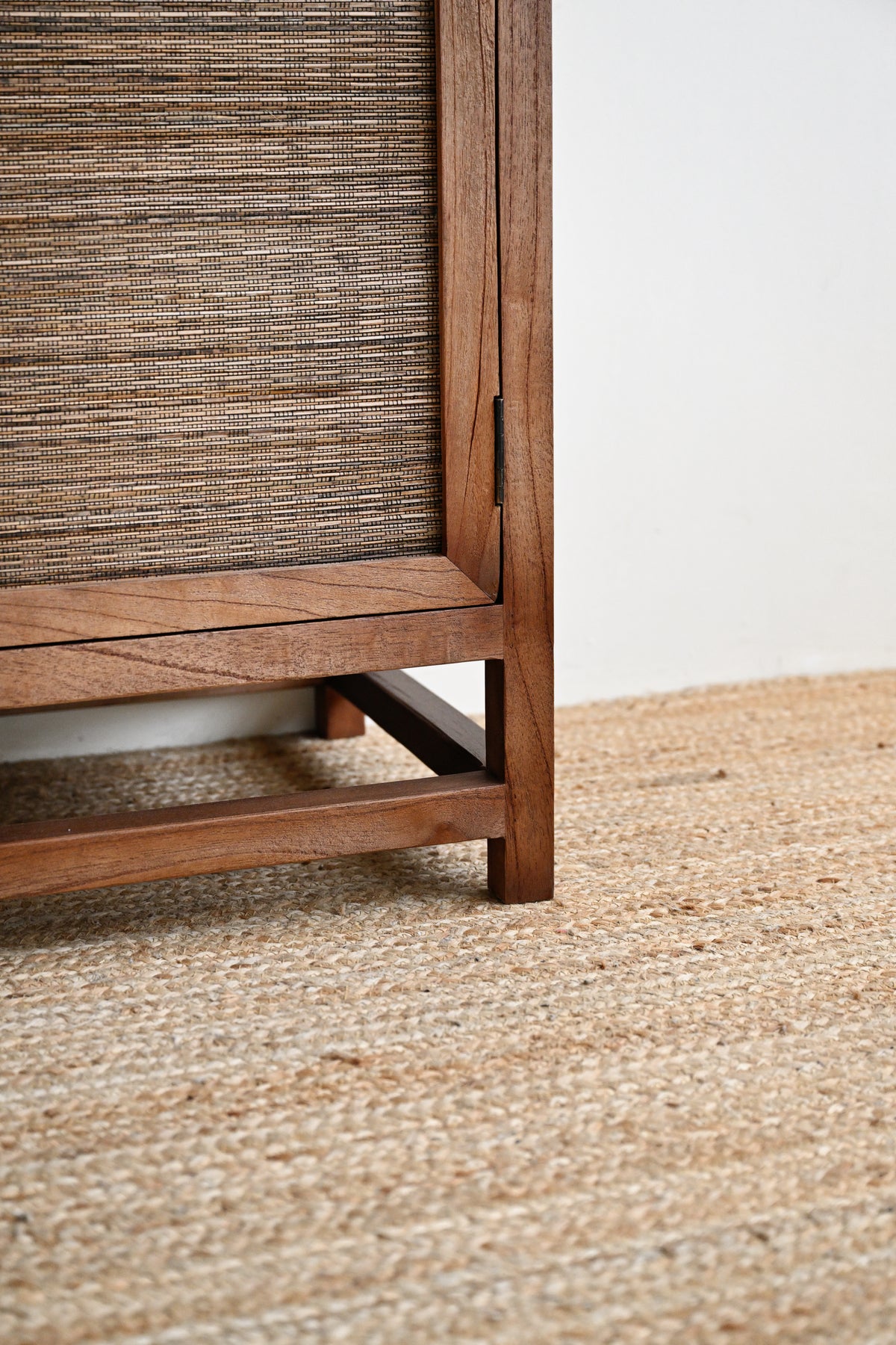 Close-up of a wooden cabinet or shelf with a woven cane or jute panel, shown standing on a natural fiber rug against a plain white wall.