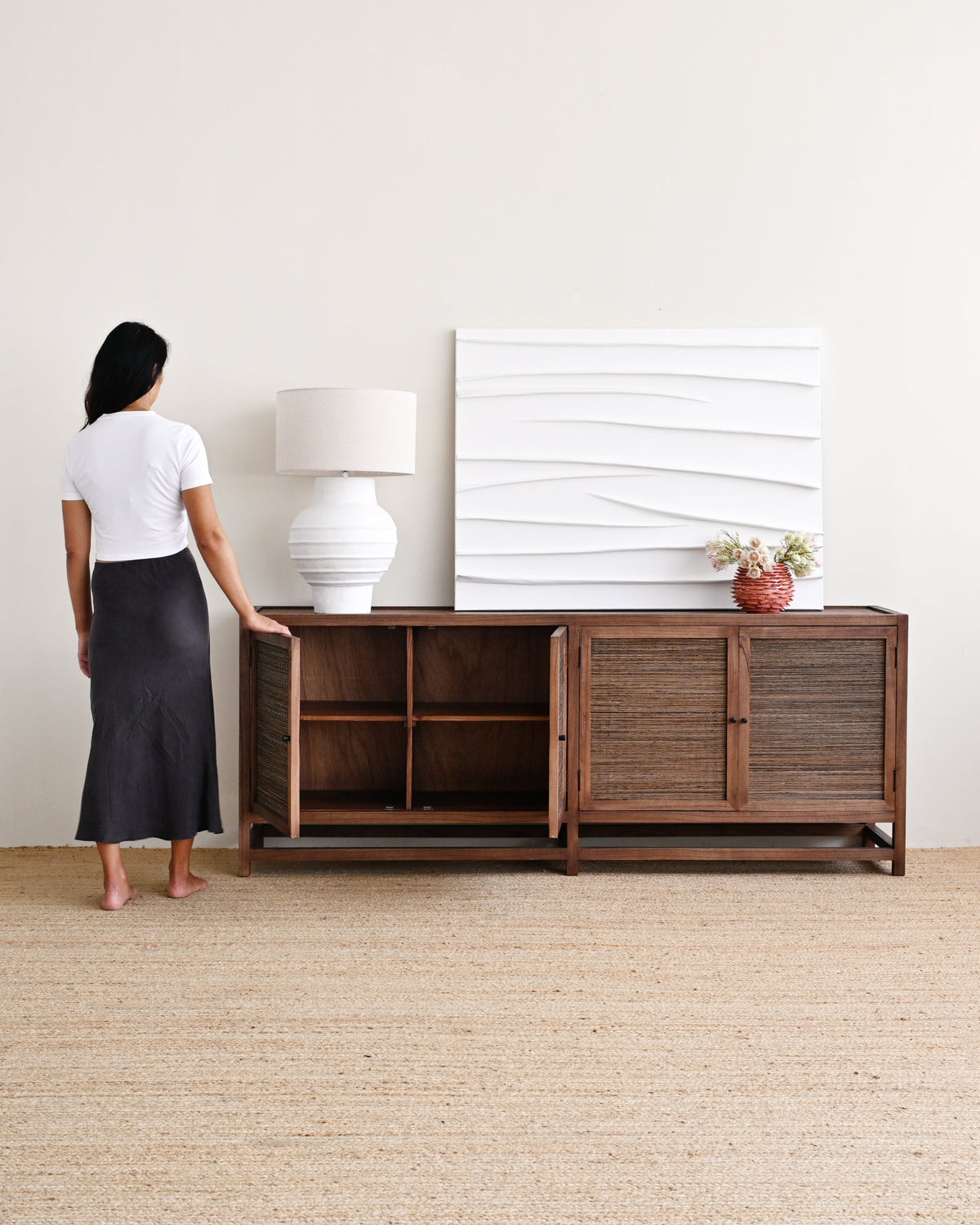 A woman stands barefoot beside a dark brown wooden sideboard or credenza in a bright, minimalist room with a white wall and textured beige rug. The cabinet has open shelving on the left and two slatted doors on the right, styled with a white ceramic lamp, a white textured wall art piece, and a small vase of flowers.