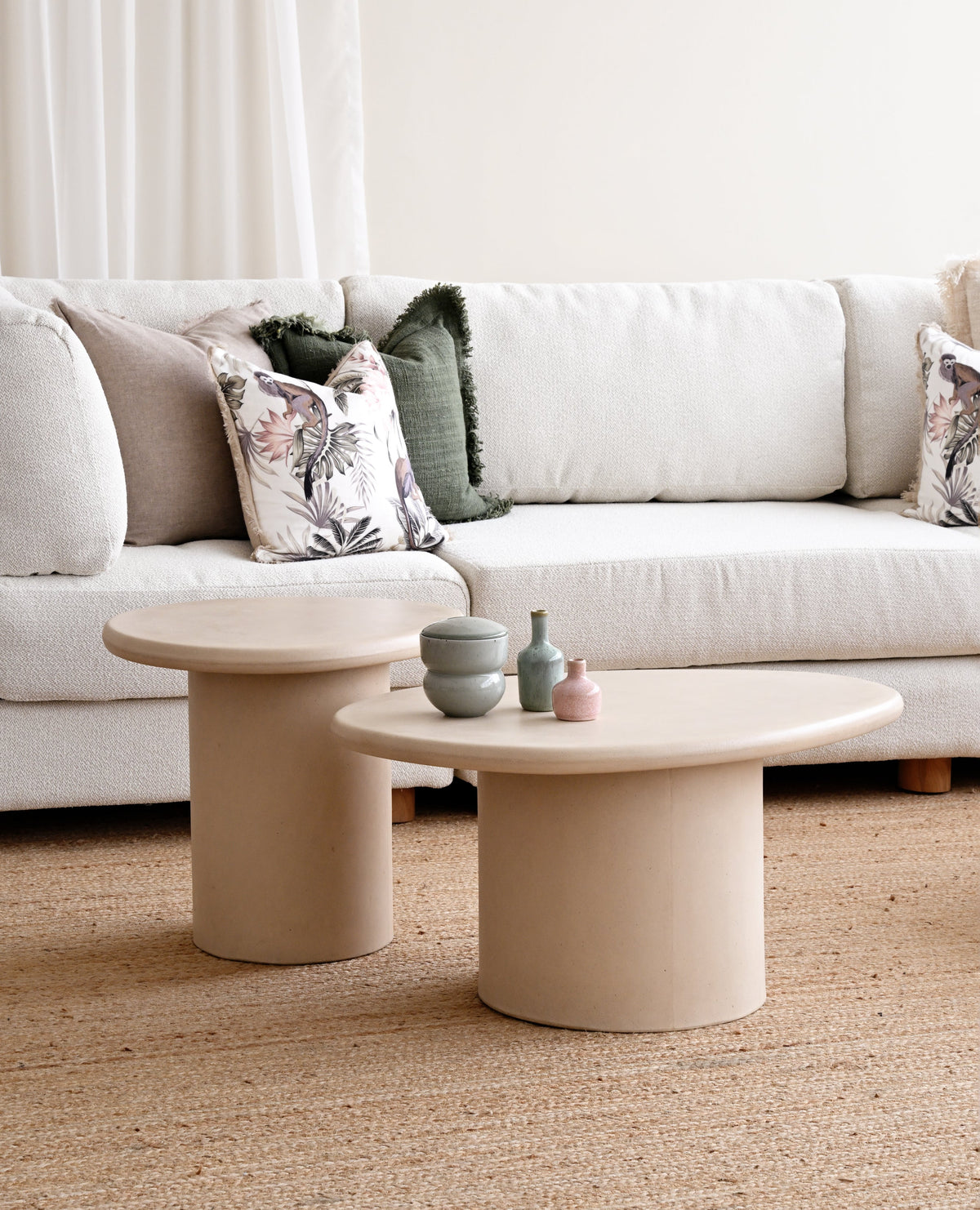A styled living room scene featuring a white fabric sofa with decorative pillows, two round beige concrete coffee tables, and a textured beige rug on the floor.