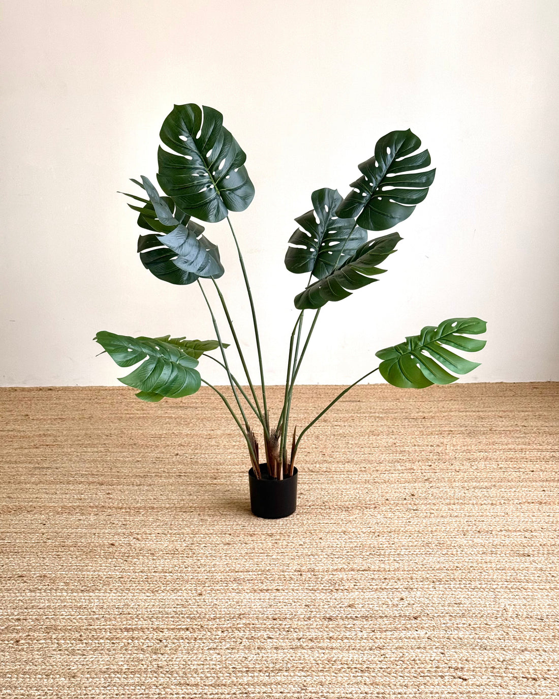 A potted artificial monstera plant with realistic green leaves and brown stems, set in a black pot on a beige woven rug against a cream wall.