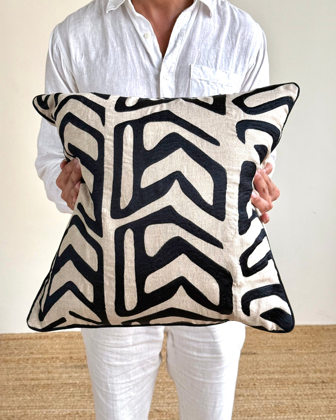 A person in a white linen shirt and white pants holds a square throw pillow with a bold black and cream geometric pattern.