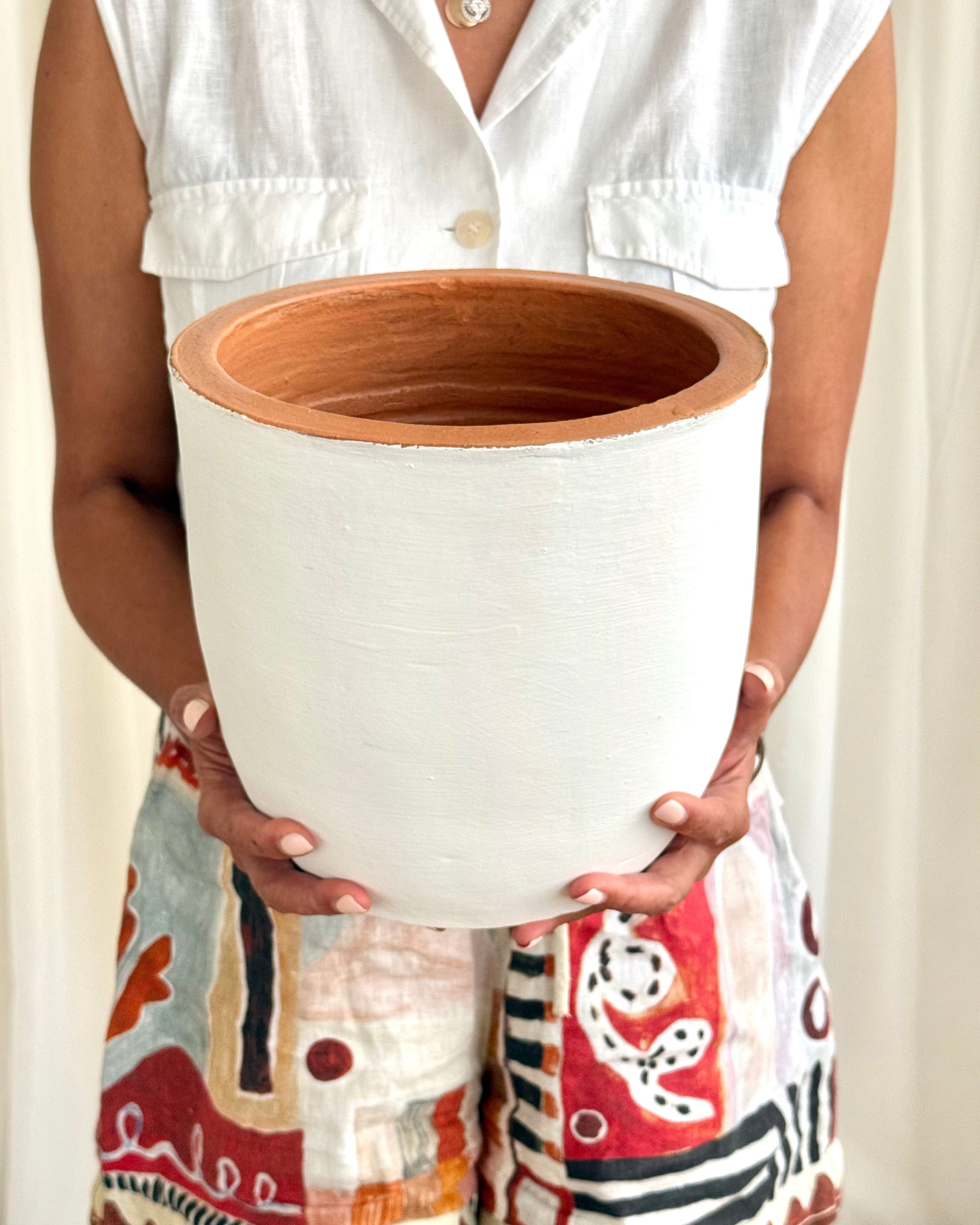 A person holding a large cylindrical white planter pot with a terracotta-colored interior, wearing a white sleeveless top and patterned shorts.