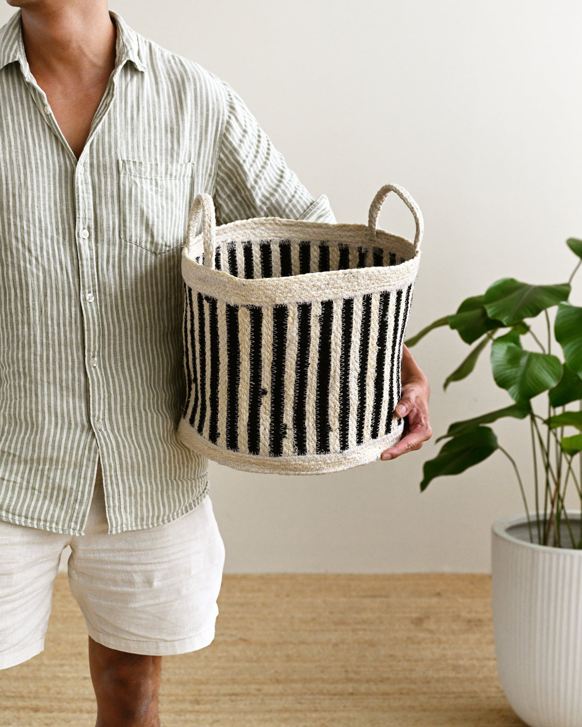 A person holding a black and cream striped woven storage basket with two loop handles, standing in a bright room with a potted plant on a light wood floor.
