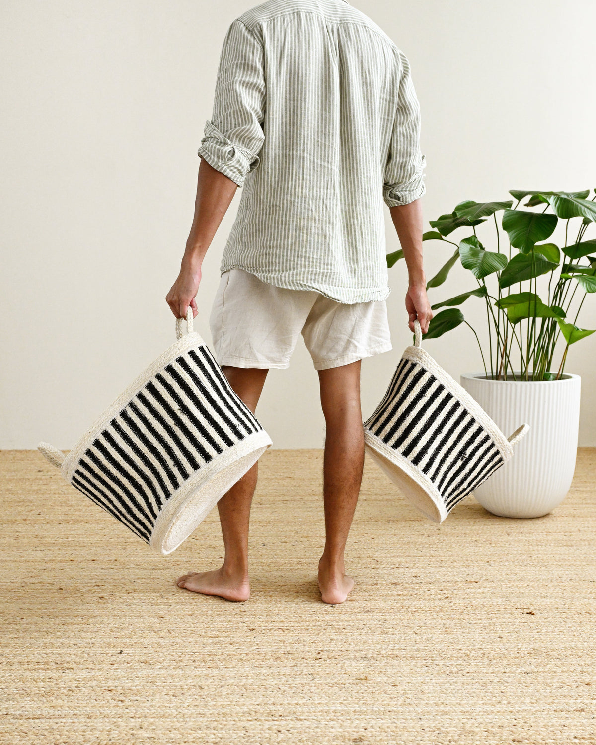 A man stands barefoot indoors holding two black-and-white striped fabric storage bins. He wears a light striped shirt and white shorts, with a potted green plant and a woven rug in the background.