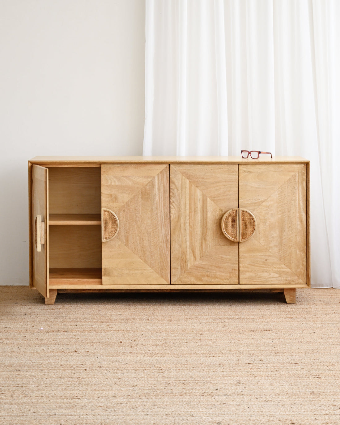 A light natural wood sideboard cabinet with geometric carved doors and circular handles, shown in a minimalist room with a white curtain and beige woven rug.