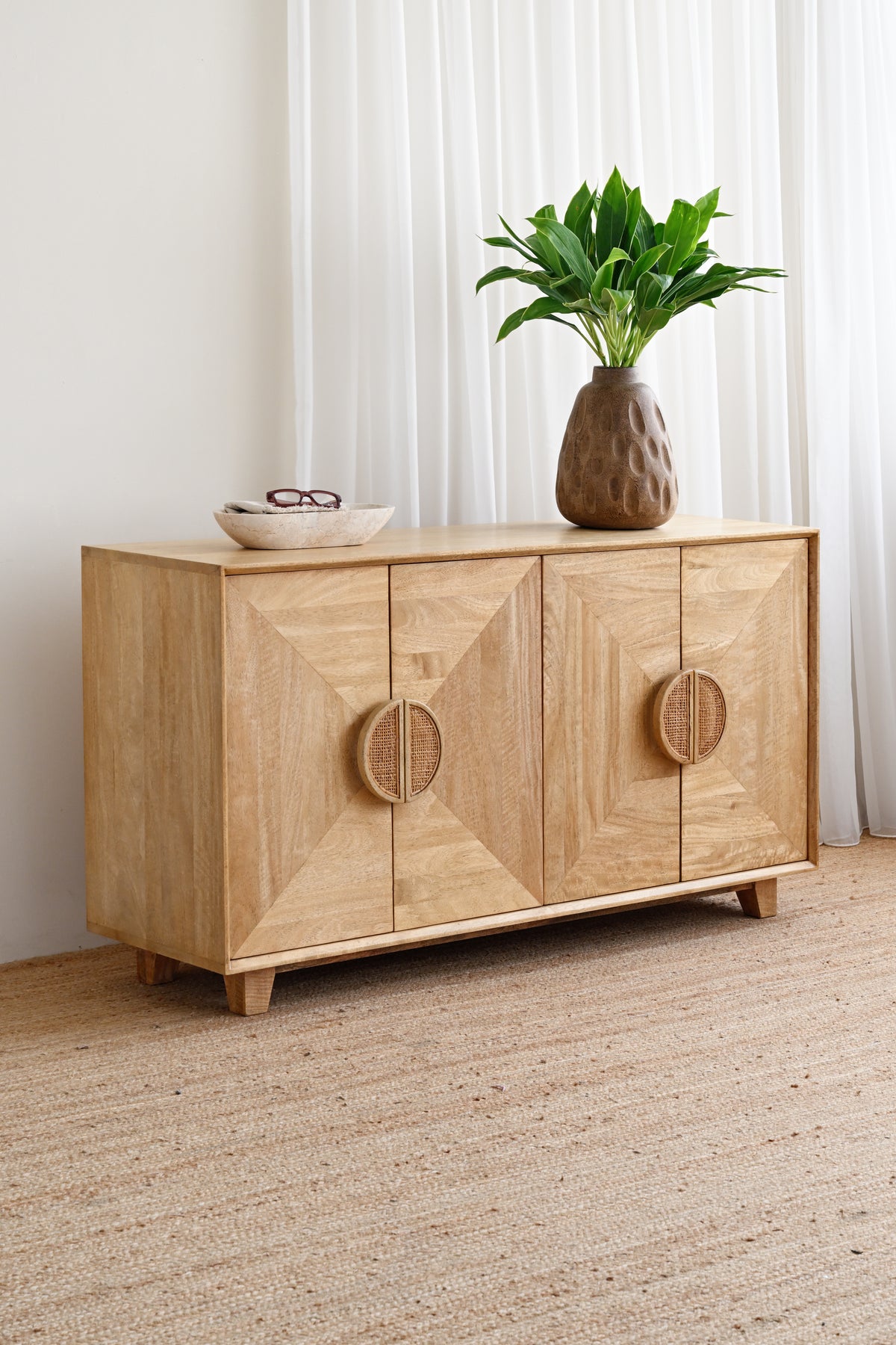 A light natural wood sideboard cabinet with geometric door panels and circular carved handles, styled in a bright room with white curtains, a woven rug, and a potted green plant on top.