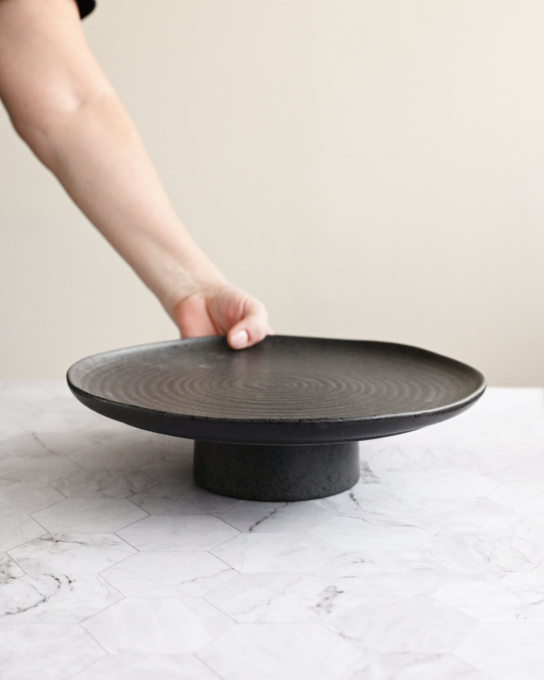 Person holding a black ceramic plate on a matching stand against a neutral background