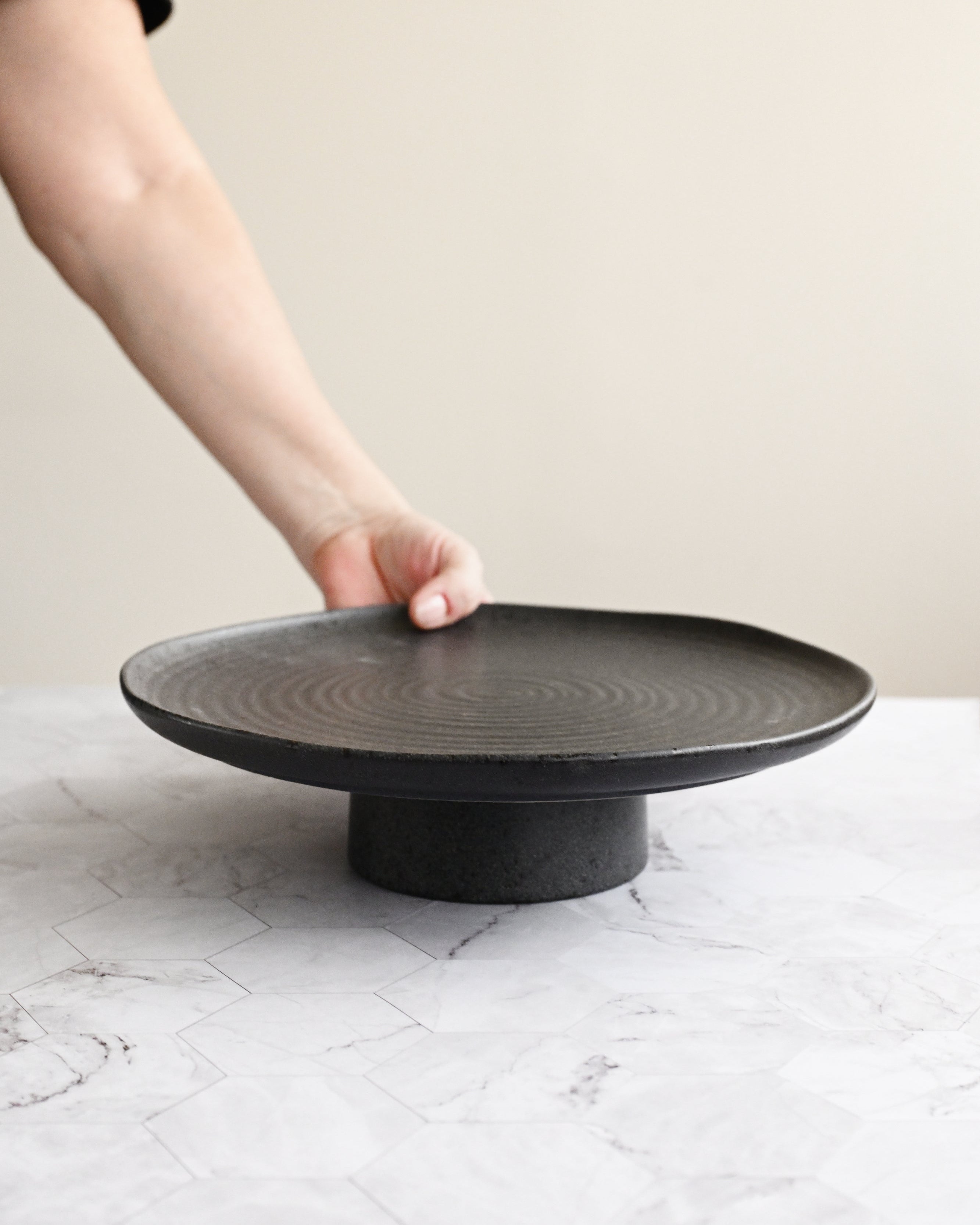 Person holding a black ceramic plate on a matching stand against a neutral background