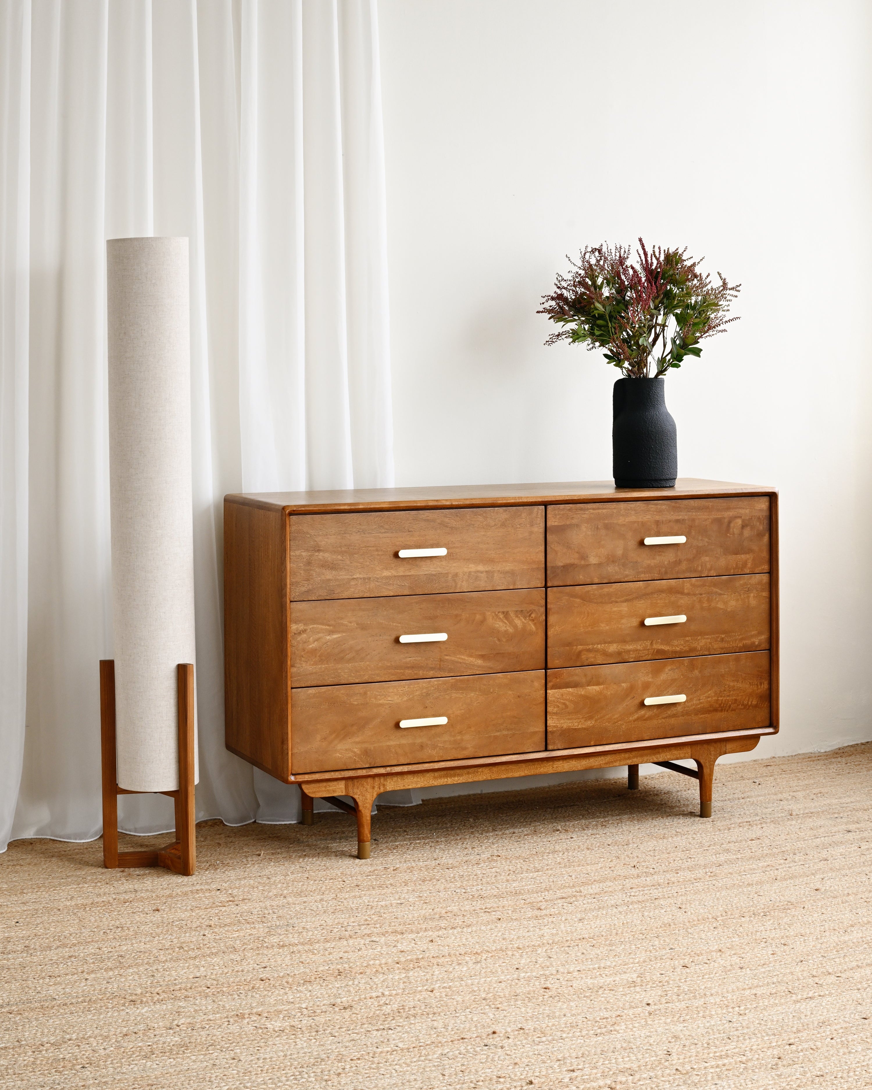 Wooden dresser with a vase of flowers next to a tall floor lamp in a room with white curtains and beige carpet.