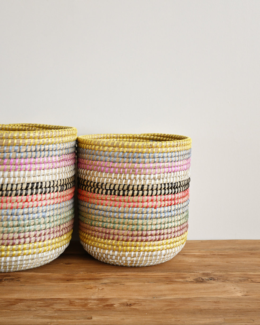 Two woven storage baskets with horizontal multicolor stripes in yellow, pink, orange, black, and white, sitting on a wooden surface against a plain light gray wall.