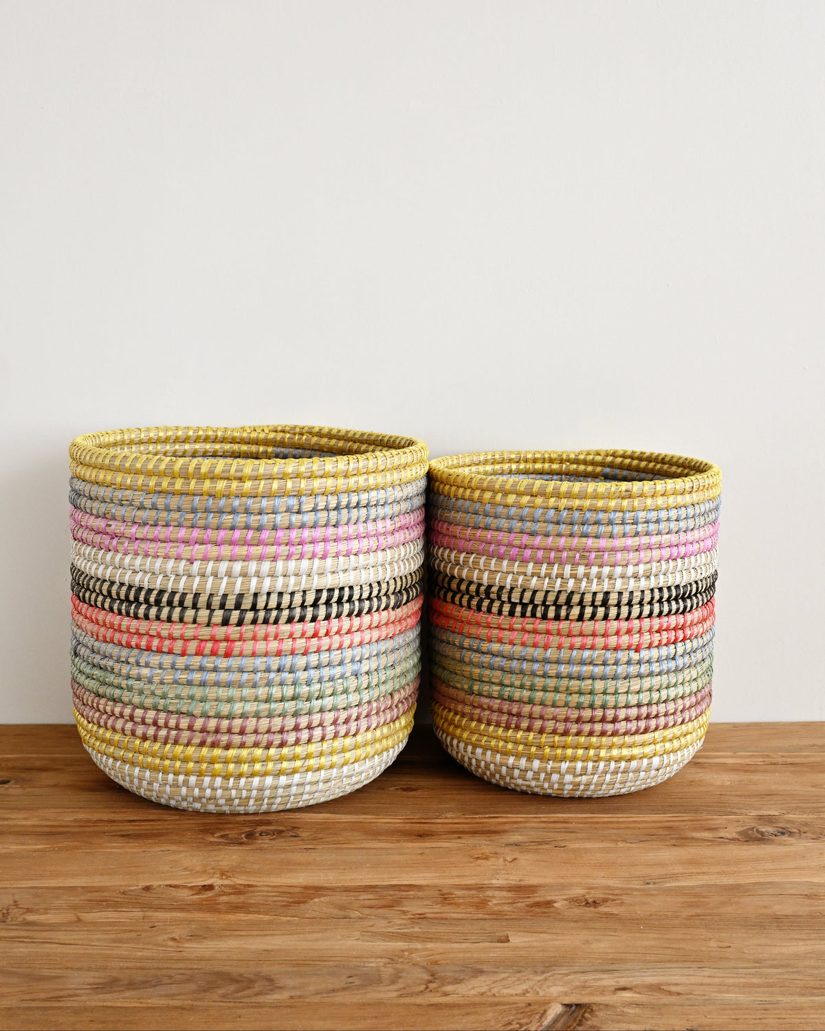 Two handwoven cylindrical baskets with horizontal multicolor stripes in yellow, pink, blue, black, and white, displayed on a wooden surface against a plain white wall.