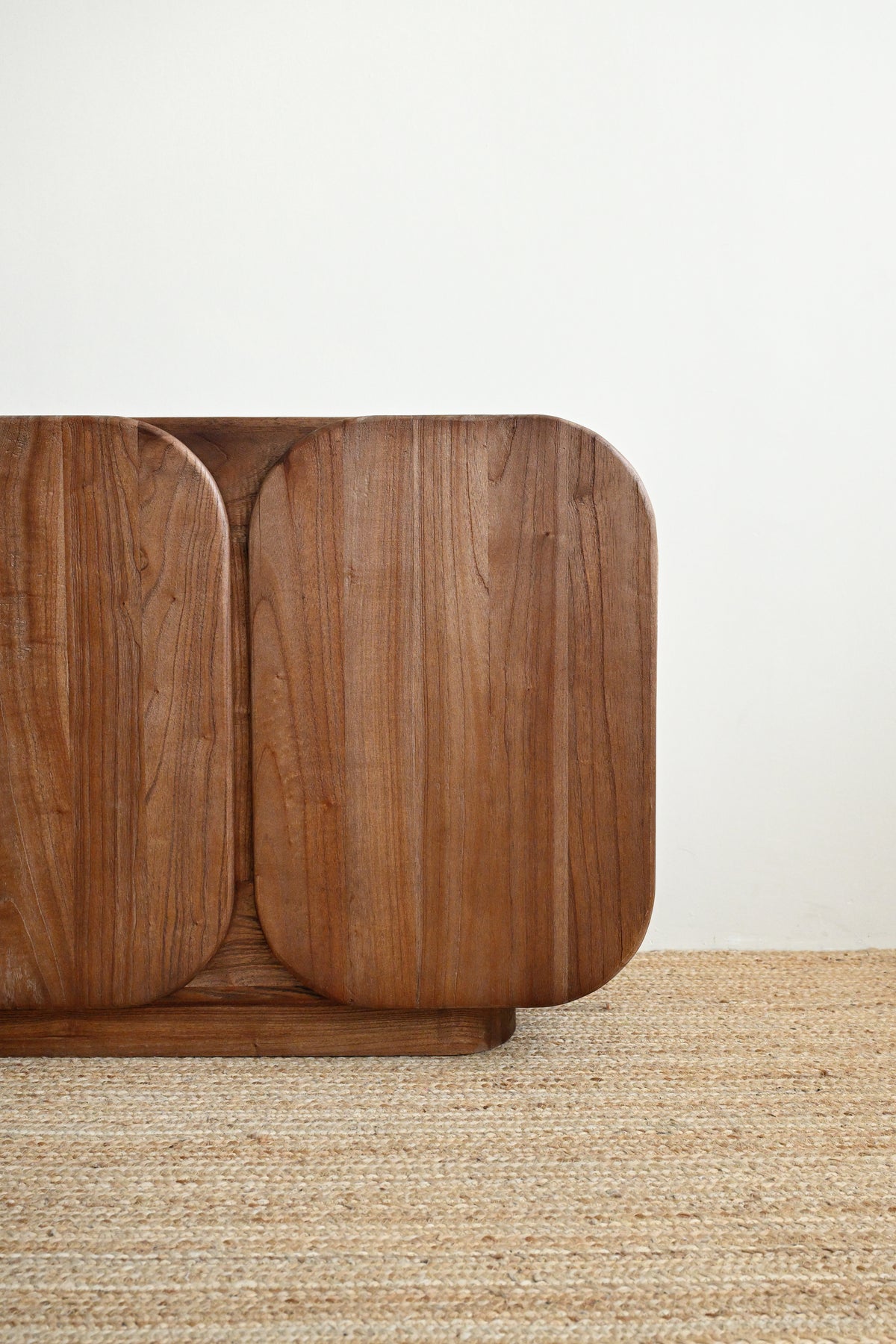 A close-up of a mid-century modern wooden cabinet with rounded edges and a warm walnut finish, set against a plain white wall and a textured beige woven rug.