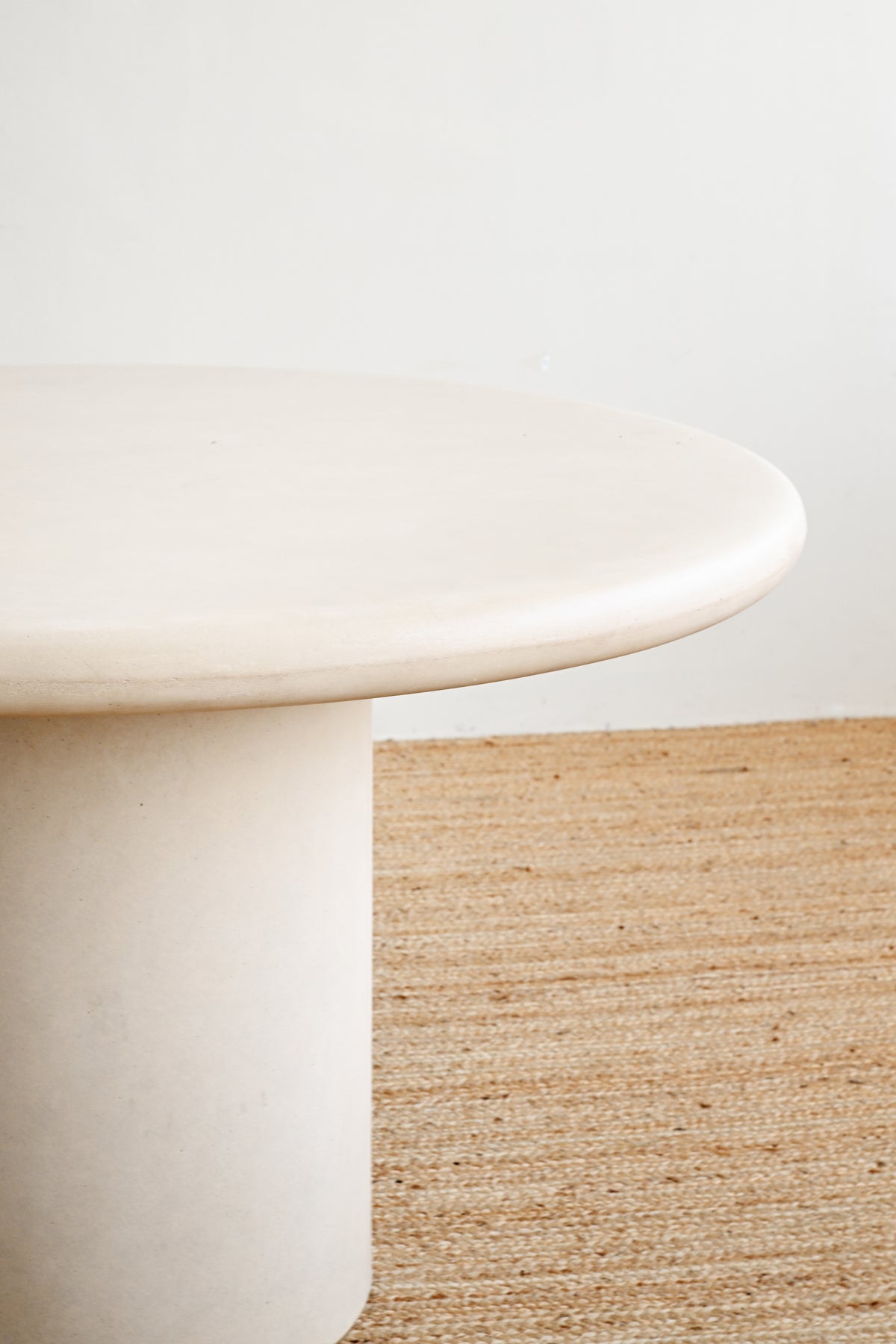 A close-up of a round, low-profile table with a smooth, matte off-white finish, set against a neutral wall and a woven natural fiber rug.