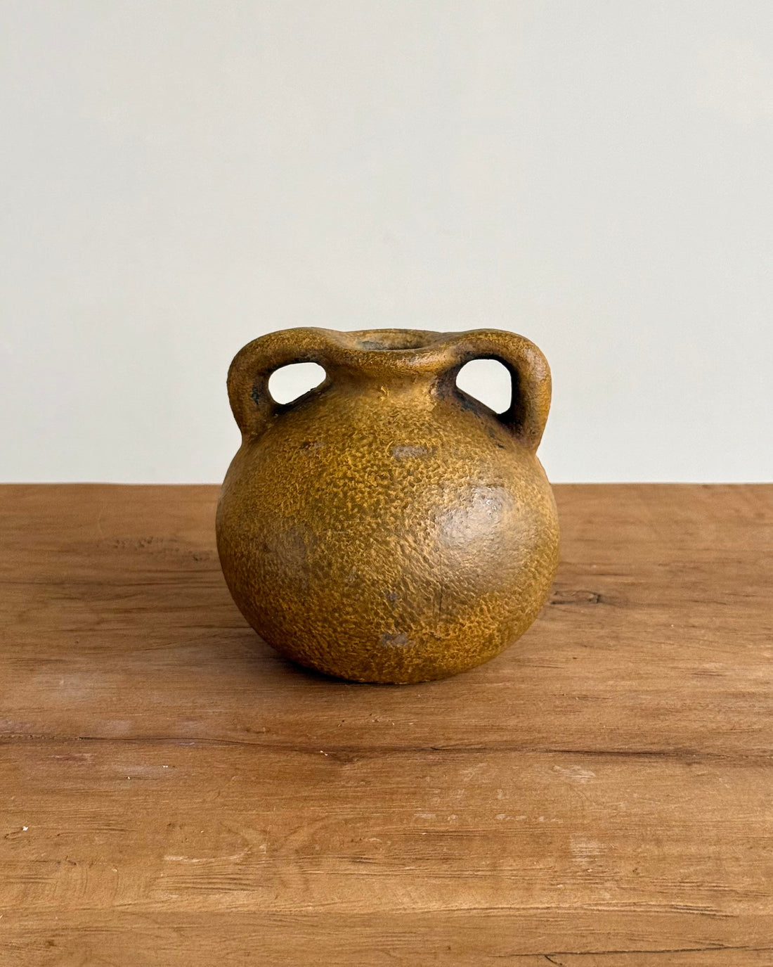 A small round ceramic vase with two loop handles, featuring a textured speckled glaze in earthy brown and ochre tones, set on a wooden tabletop against a plain light gray wall.