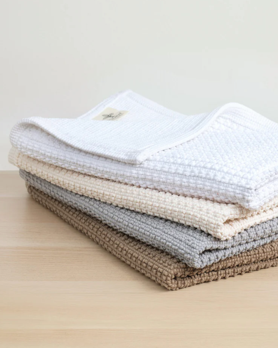 A stack of folded Heveya vegan cotton bath mats in beige, gray, and brown colors, showcasing the honeycomb texture.