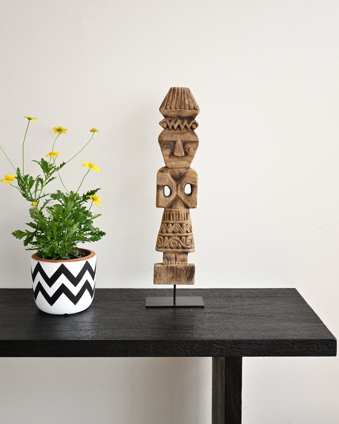 Wooden totem figure on a black table with a potted plant against a white wall