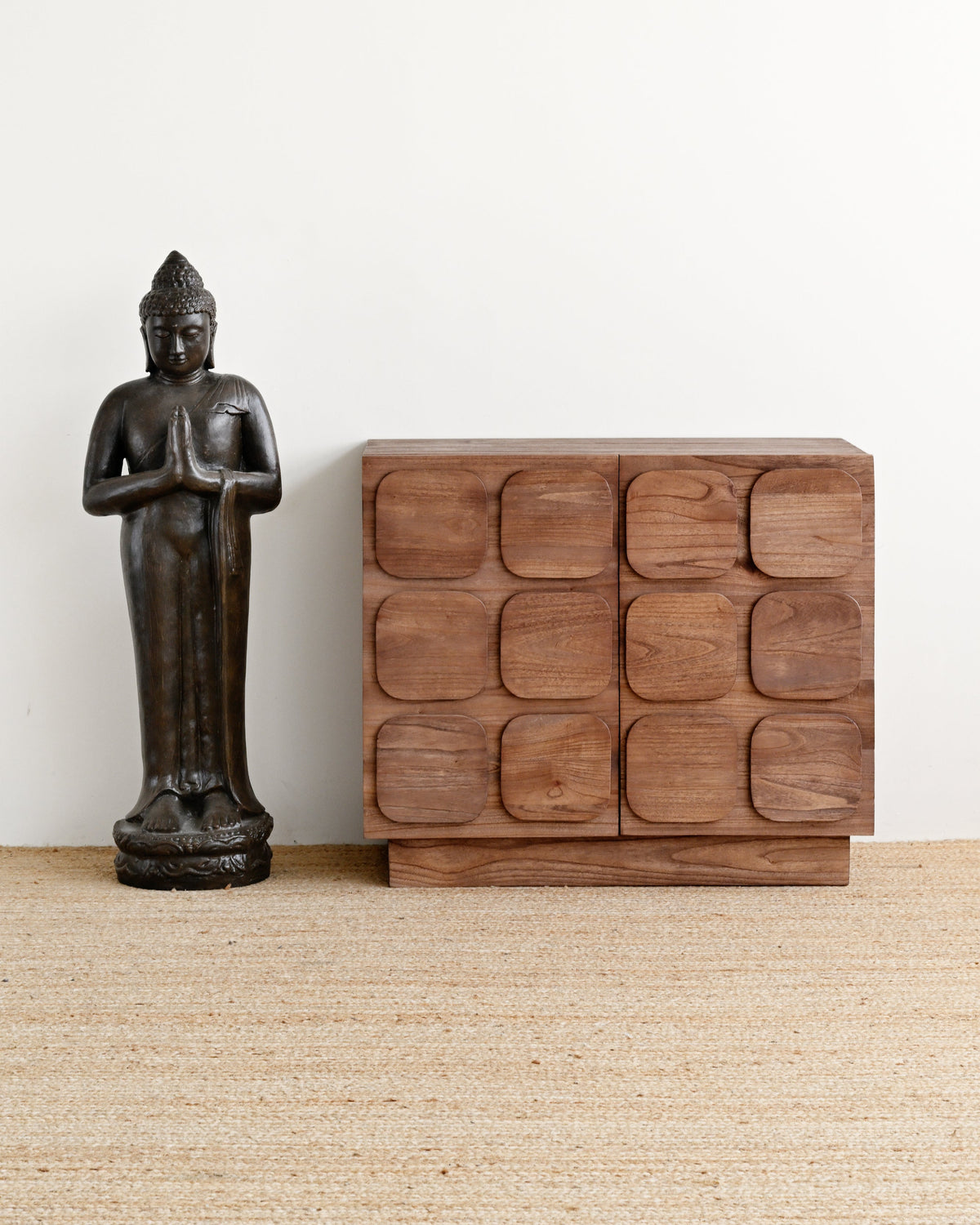 A dark bronze standing Buddha statue next to a mid-century modern wooden sideboard with a wavy geometric front, set on a woven jute rug against a plain white wall.