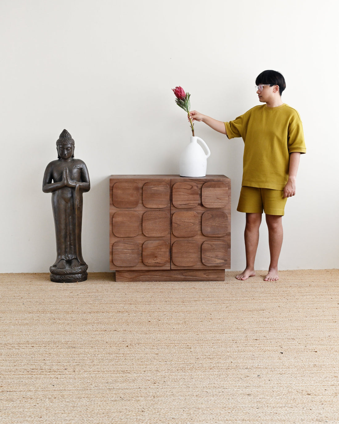 A child wearing a mustard yellow t-shirt and matching shorts stands barefoot next to a carved wooden chest, holding a white vase with a red flower, in a minimalist room with a Buddha statue and a woven rug.