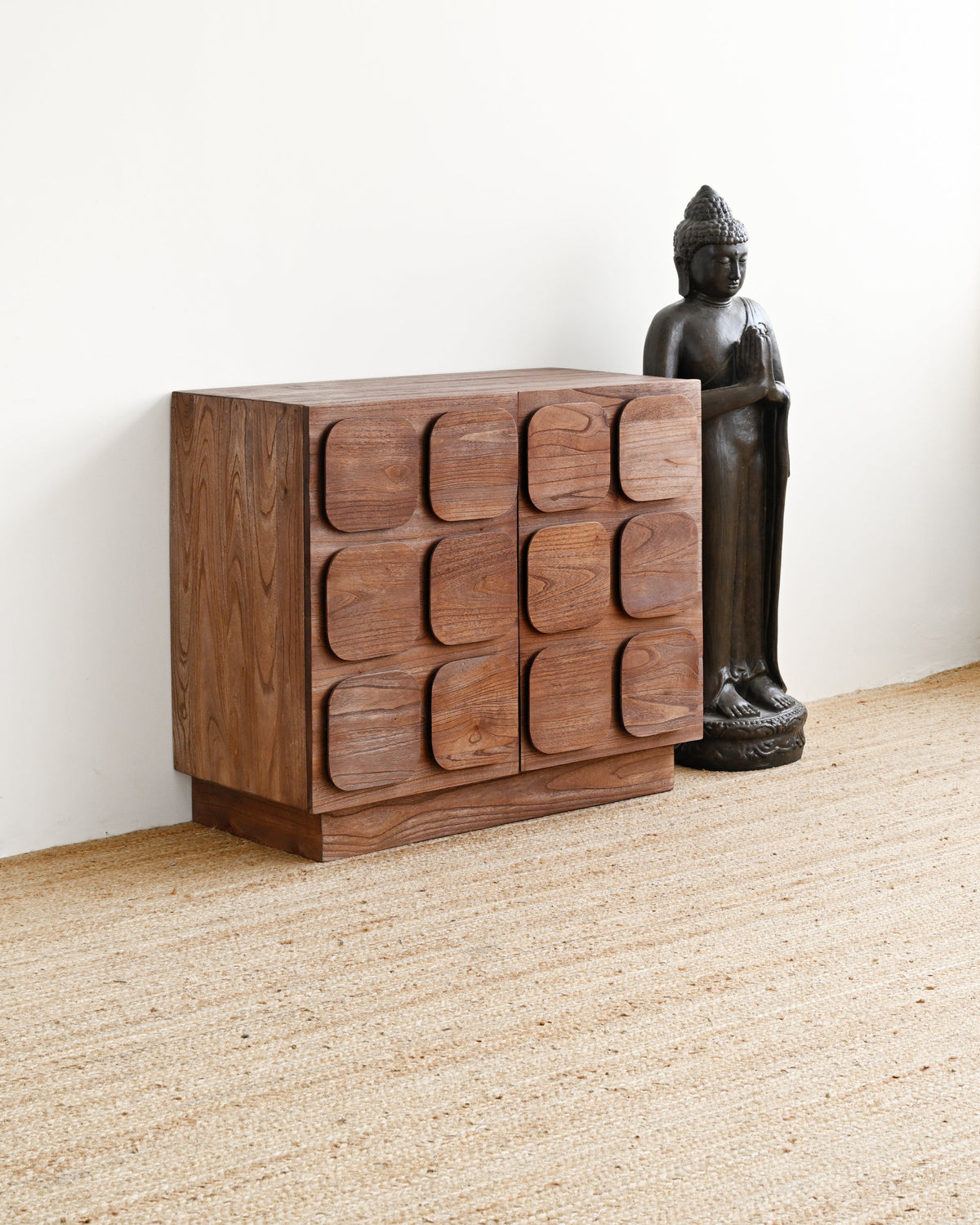 A dark brown wooden chest of drawers with a modern geometric, wavy front design, styled next to a black Buddha statue on a beige woven rug against a white wall.