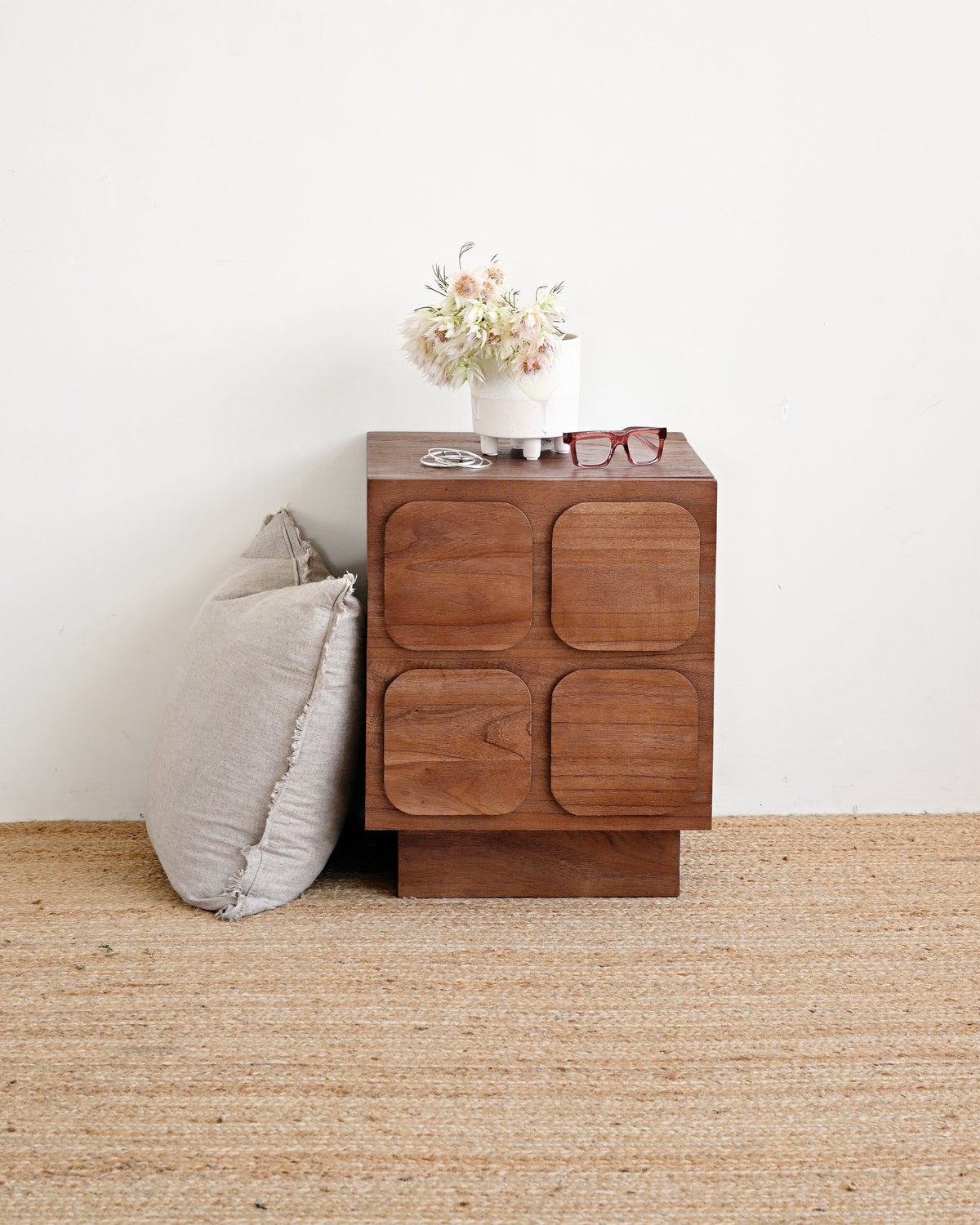 A mid-century modern wooden nightstand with four square recessed drawer fronts, styled next to a gray pillow on a woven jute rug against a white wall.