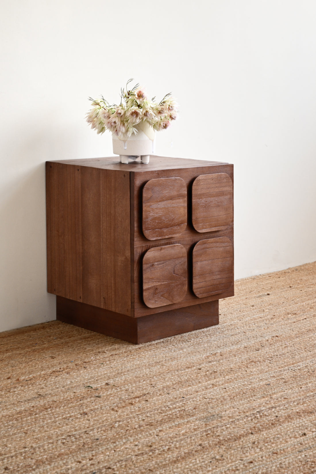 A mid-century modern wooden side table with four rounded square drawer fronts, shown on a woven jute rug against a plain off-white wall with a vase of flowers on top.