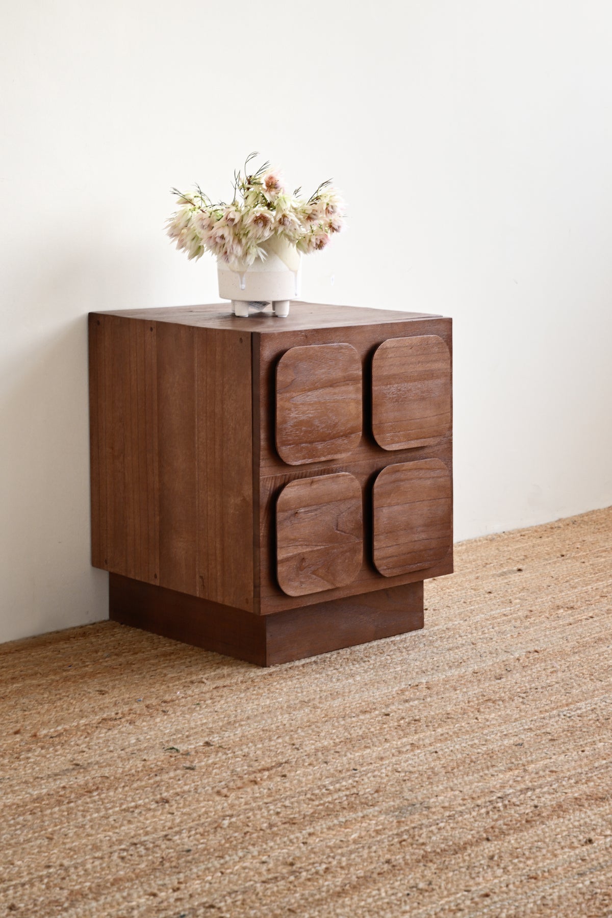 A mid-century modern wooden side table with four rounded square drawer fronts, shown on a woven jute rug against a plain off-white wall with a vase of flowers on top.