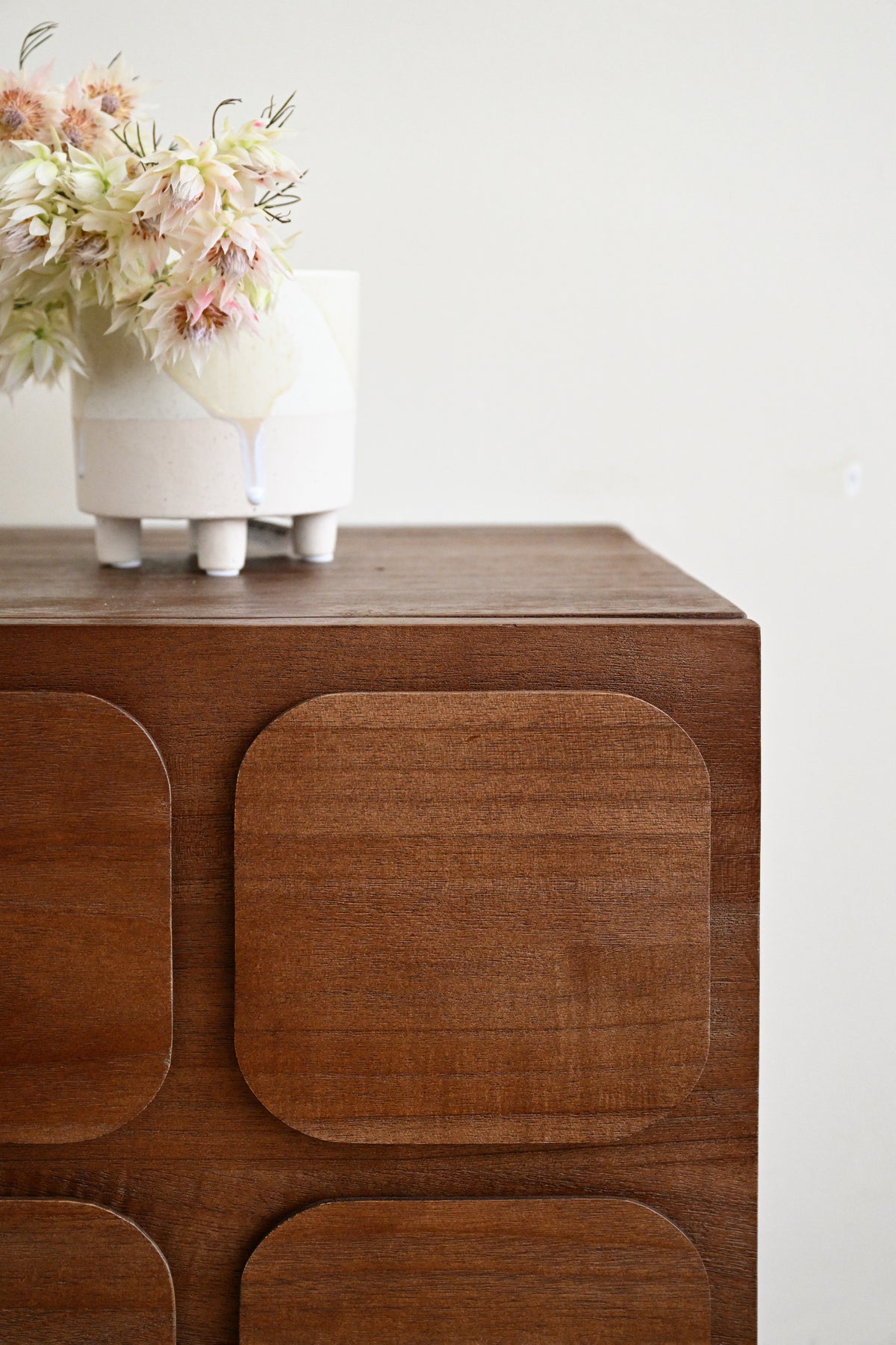 A close-up of a mid-century modern wooden cabinet with rounded square panel details on the front, styled with a white ceramic planter holding dried pink and white flowers on top, set against a plain off-white wall.