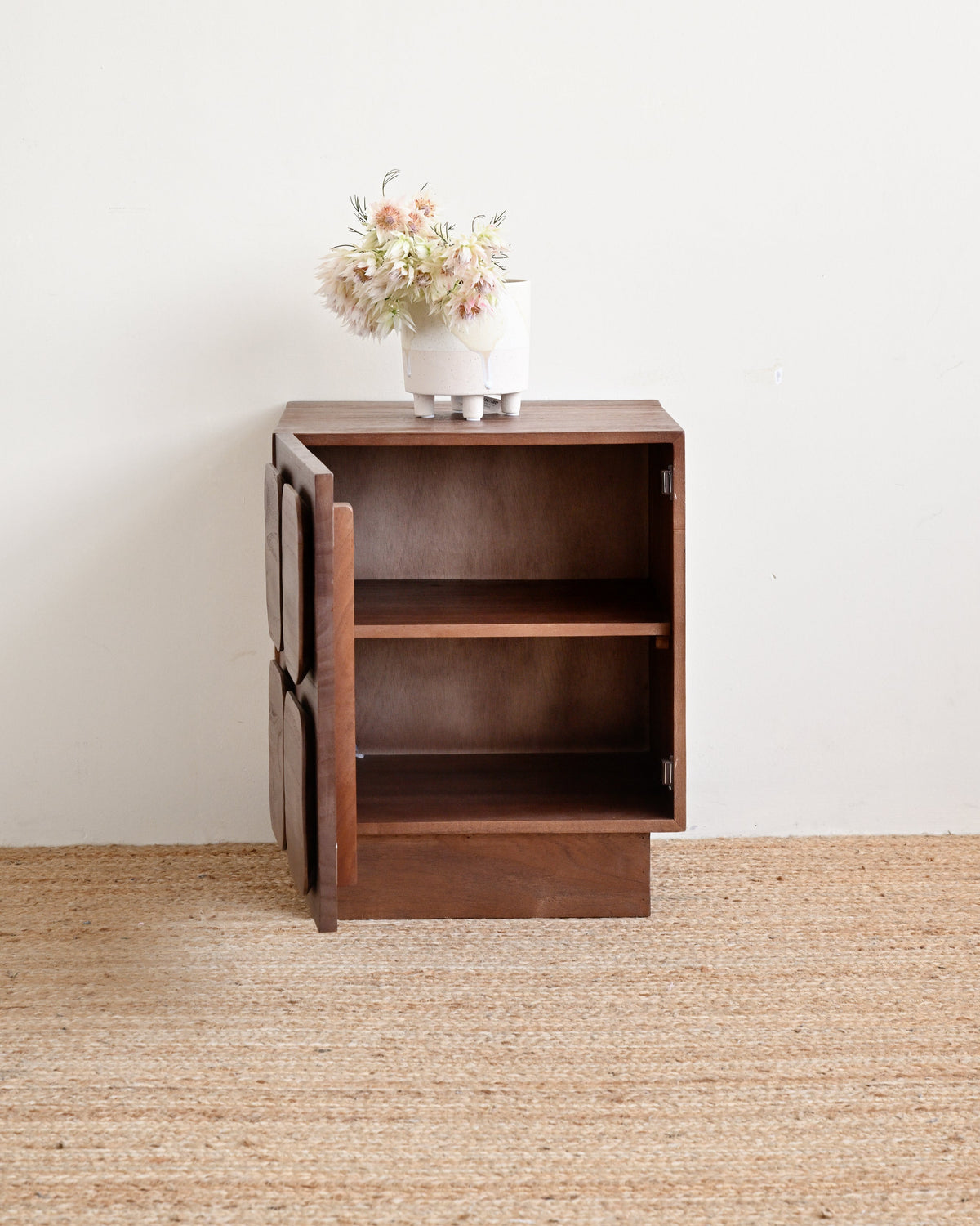 A small dark brown wooden side cabinet with one open door and two interior shelves, shown on a woven jute rug against a plain white wall. A white vase with pale pink flowers sits on top.