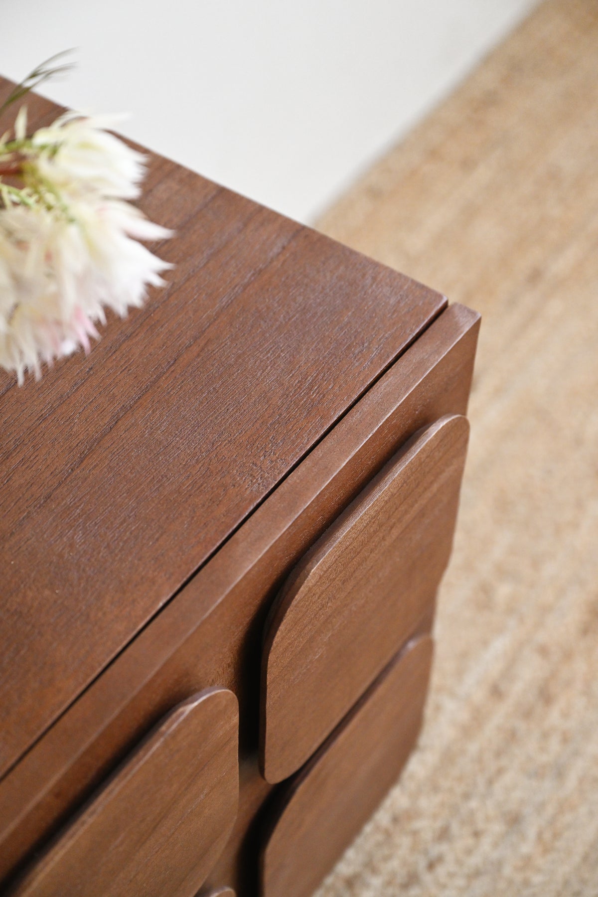 A close-up of a dark brown wooden side table or cabinet with rounded drawer fronts, shown in a styled interior setting with a beige rug and a white flower on top.