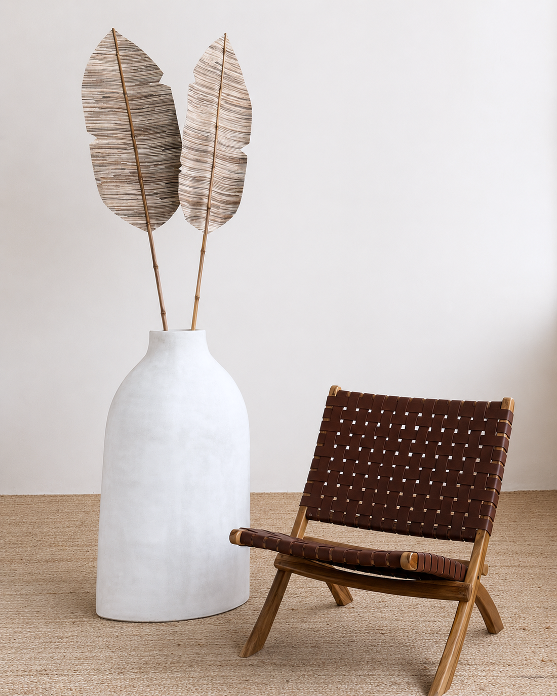 White vase with dried leaves next to a brown woven chair on a beige carpet.