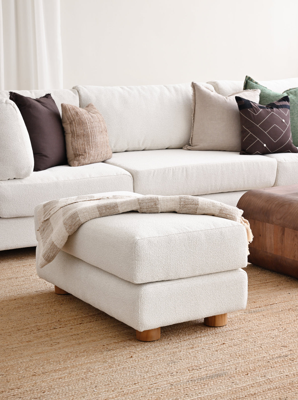 A modern two-tiered upholstered ottoman in a warm off-white bouclé fabric, styled with a beige plaid throw blanket in a bright, contemporary living room setting.