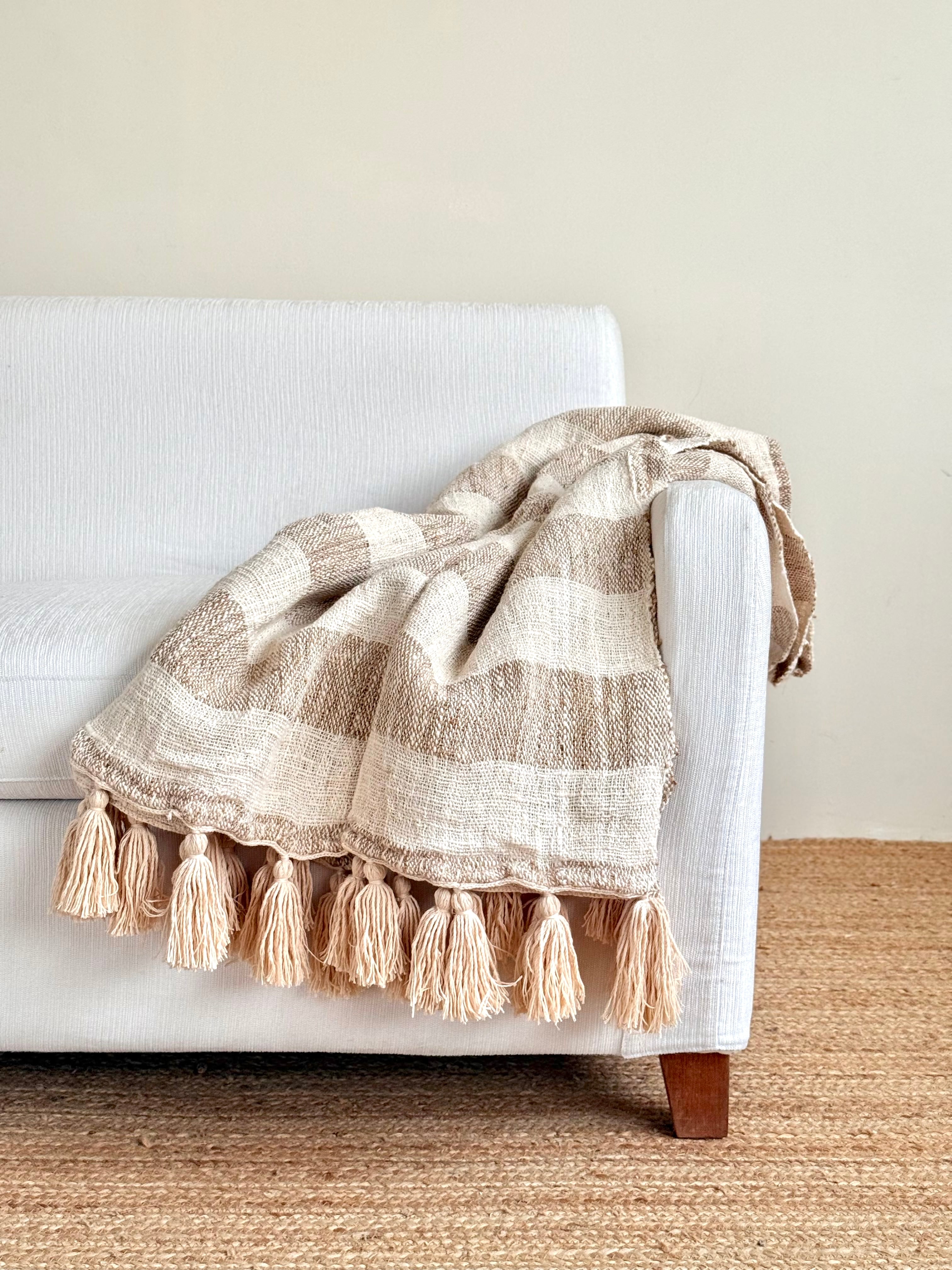 A woven throw blanket with beige and cream stripes and tassels, draped over a white armchair in a minimalist room setting.
