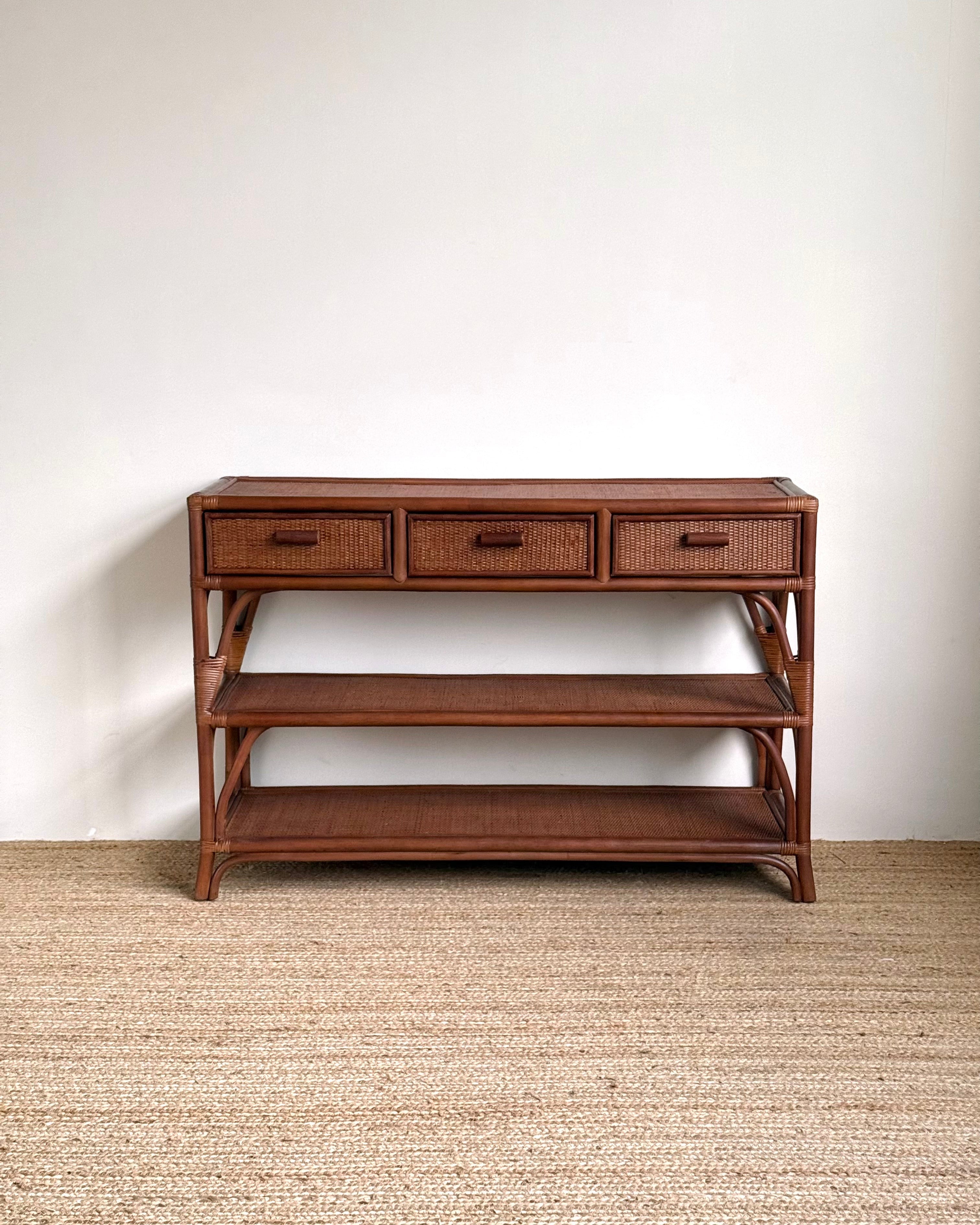 A mid-century modern wooden console table with three drawers, woven rattan drawer fronts, an open middle shelf, and a lower shelf, set against a plain white wall and light beige woven rug.