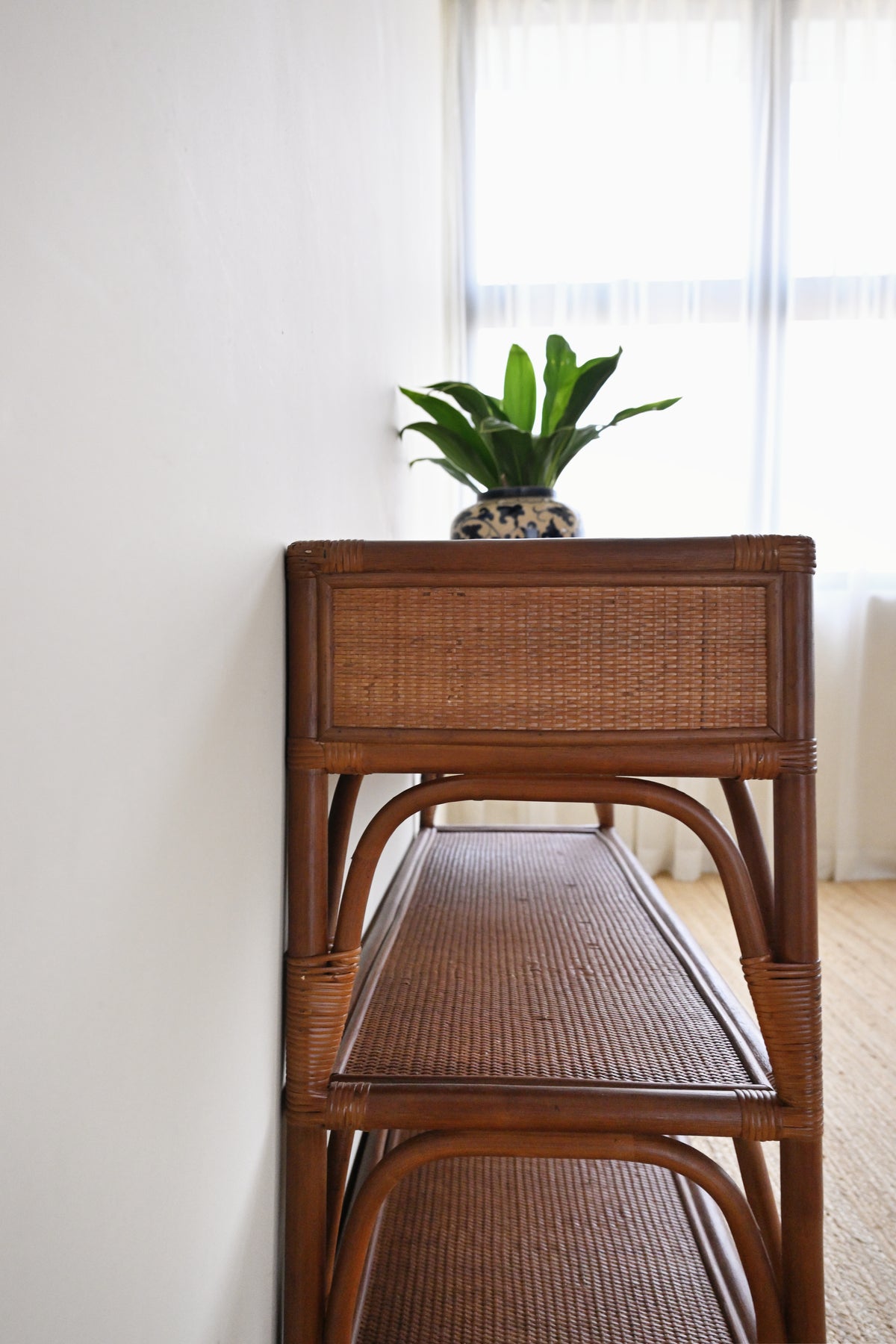 A brown wooden rattan console shelf with two tiers, styled with a potted green plant on top, set against a white wall and window background.
