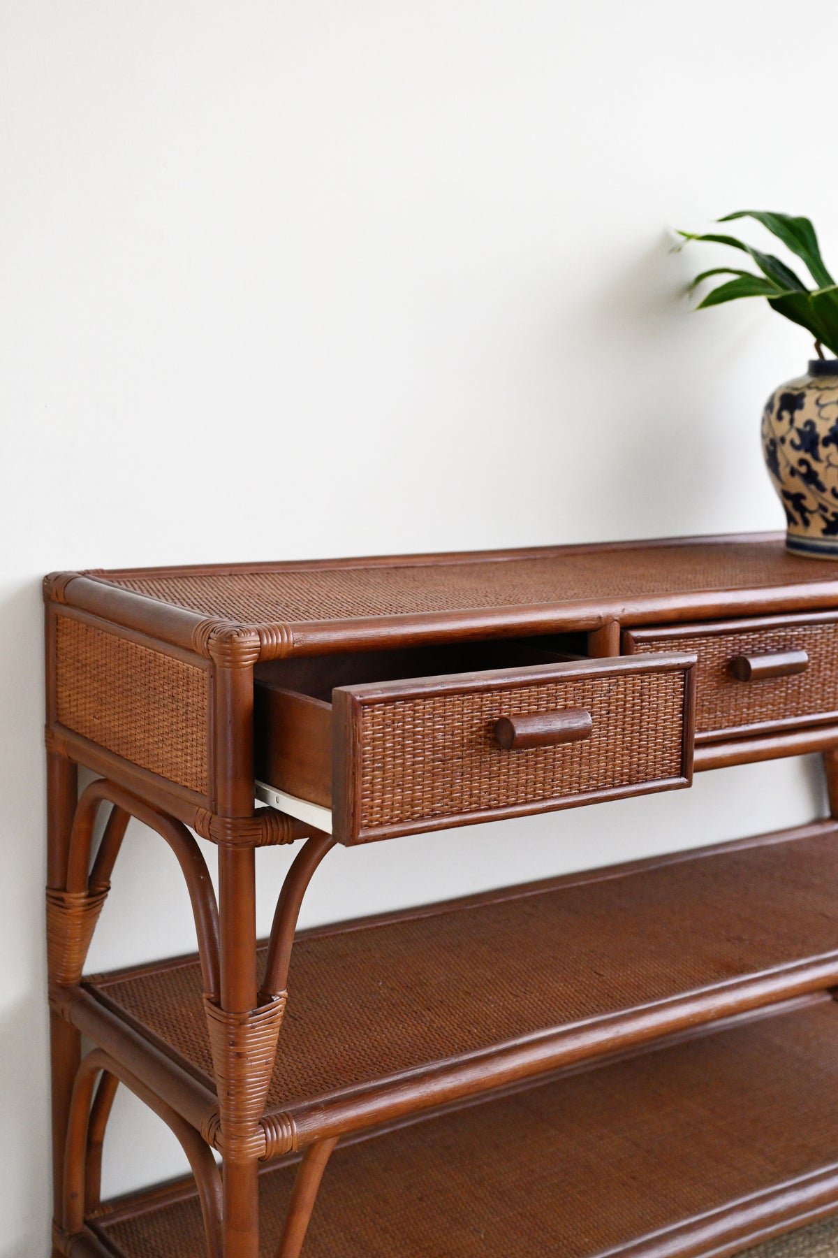 A brown rattan console table with woven drawer fronts and wooden handles, shown against a plain white wall with a potted plant in the background.
