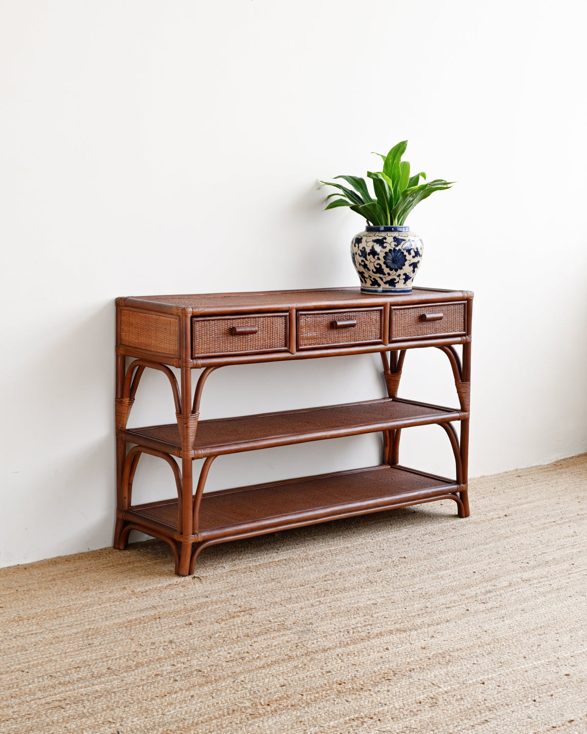 A mid-century modern wooden console table with three drawers, open shelves, and curved bamboo-style legs, styled with a potted green plant on top, set against a plain white wall and light beige woven rug.