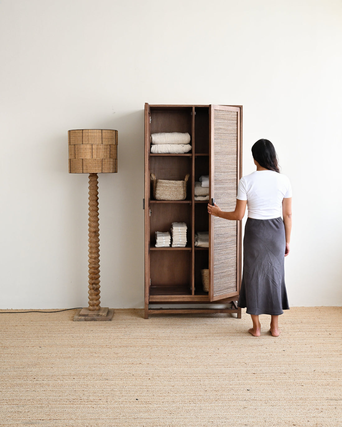 A woman stands in front of a tall wooden cabinet with slatted doors, opening one side to reveal folded white linens inside. The cabinet is placed in a bright room with a woven floor lamp beside it.
