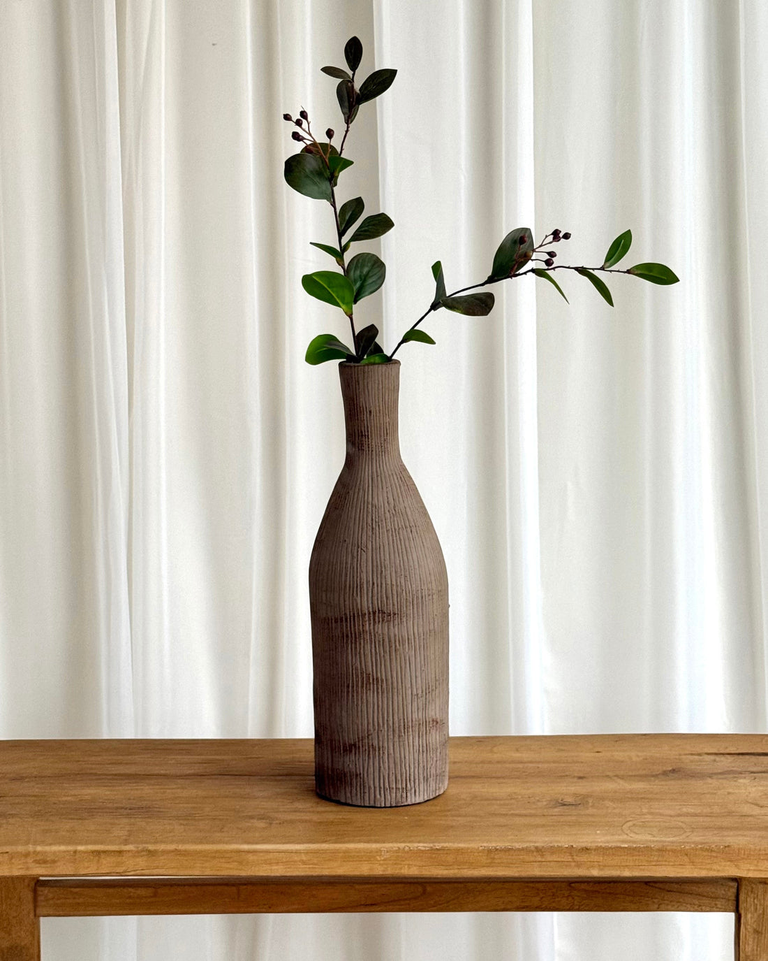 A tall ribbed ceramic vase in a warm taupe-brown color, holding a green leafy sprig, set on a rustic wooden table against a white curtain background.