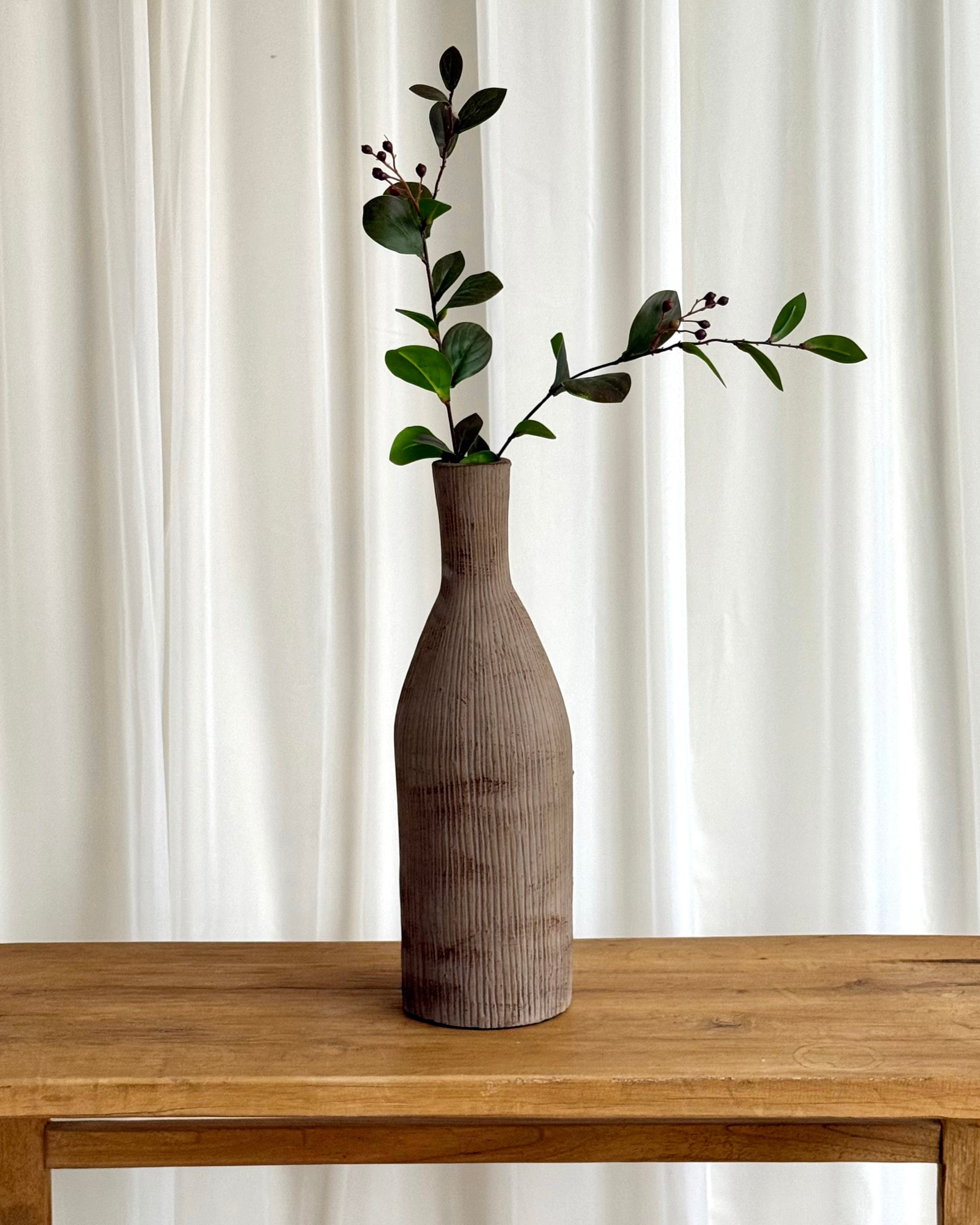 A tall ribbed ceramic vase in a warm taupe-brown color, holding a green leafy sprig, set on a rustic wooden table against a white curtain background.